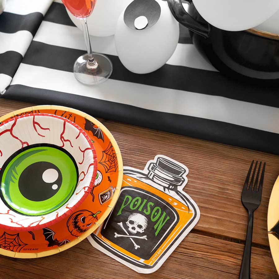 A Halloween party table setting featuring an eyeball snack bowl, black cutlery, and a die-cut paper napkin shaped like a bottle of orange poison.