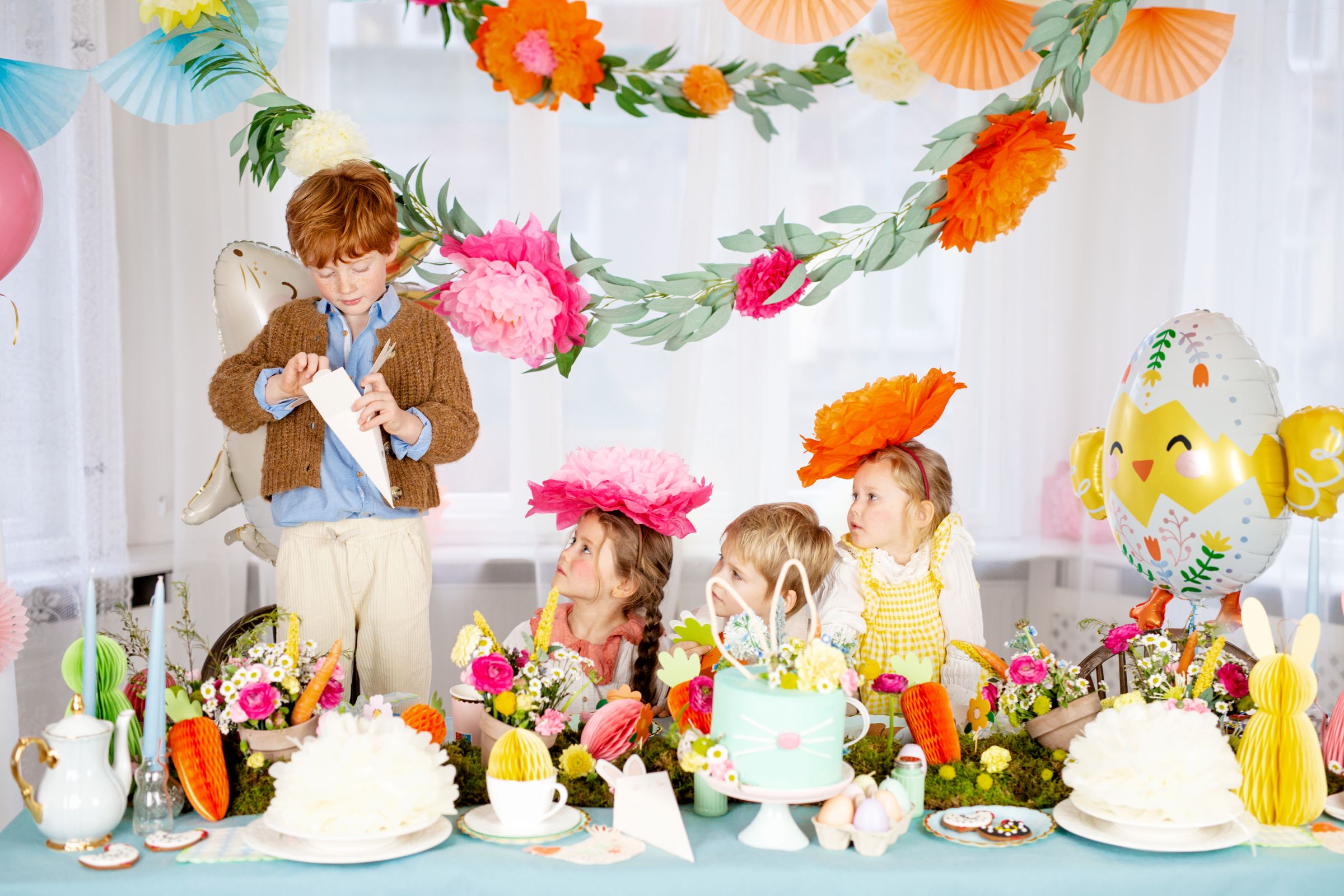 A cheerful chick in a floral eggshell foil balloon floating above a festive Easter party table decorated with flowers and treats.
