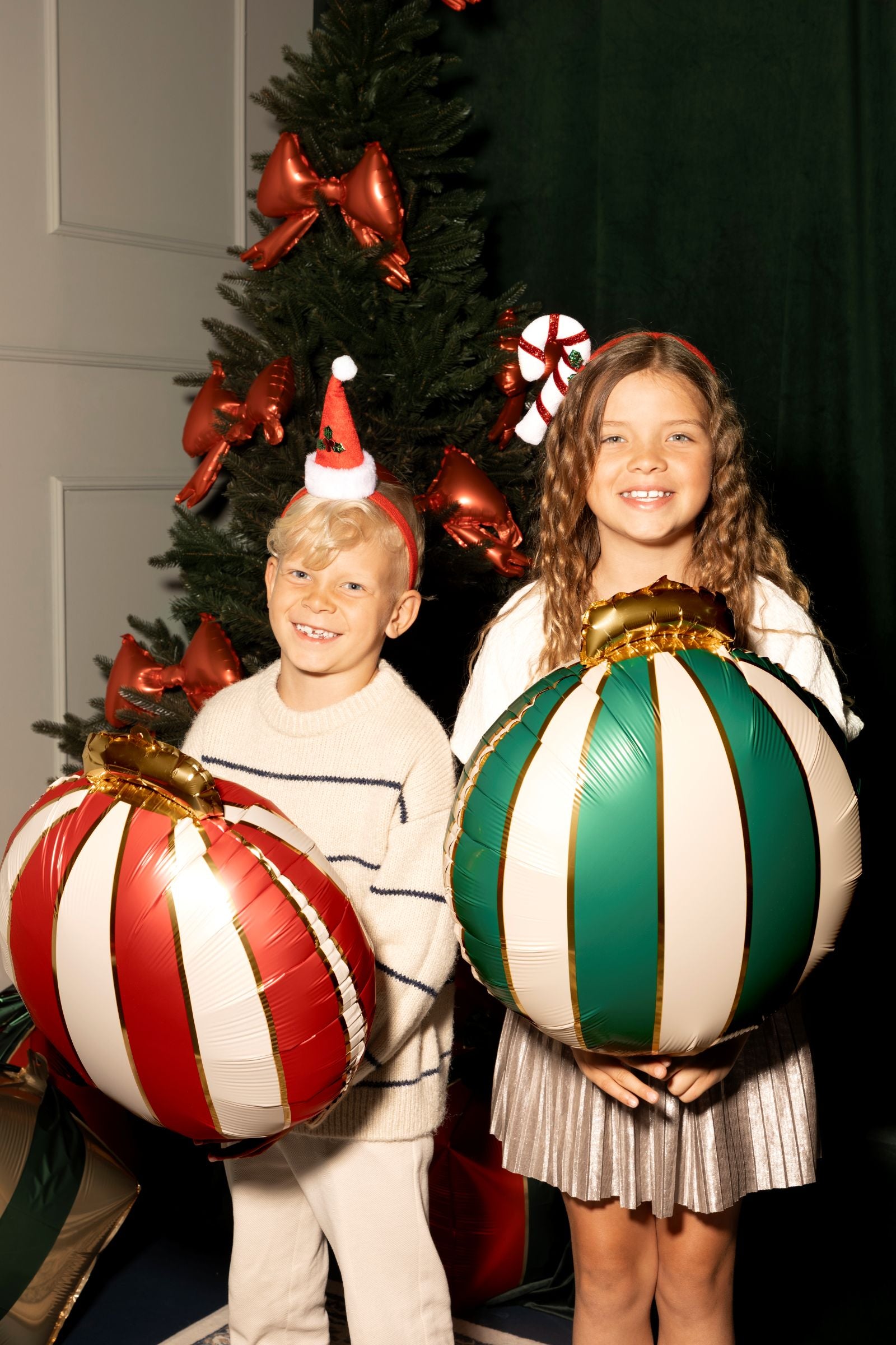 Two children smiling by a Christmas tree, each holding a large foil balloon shaped like a Christmas bauble.