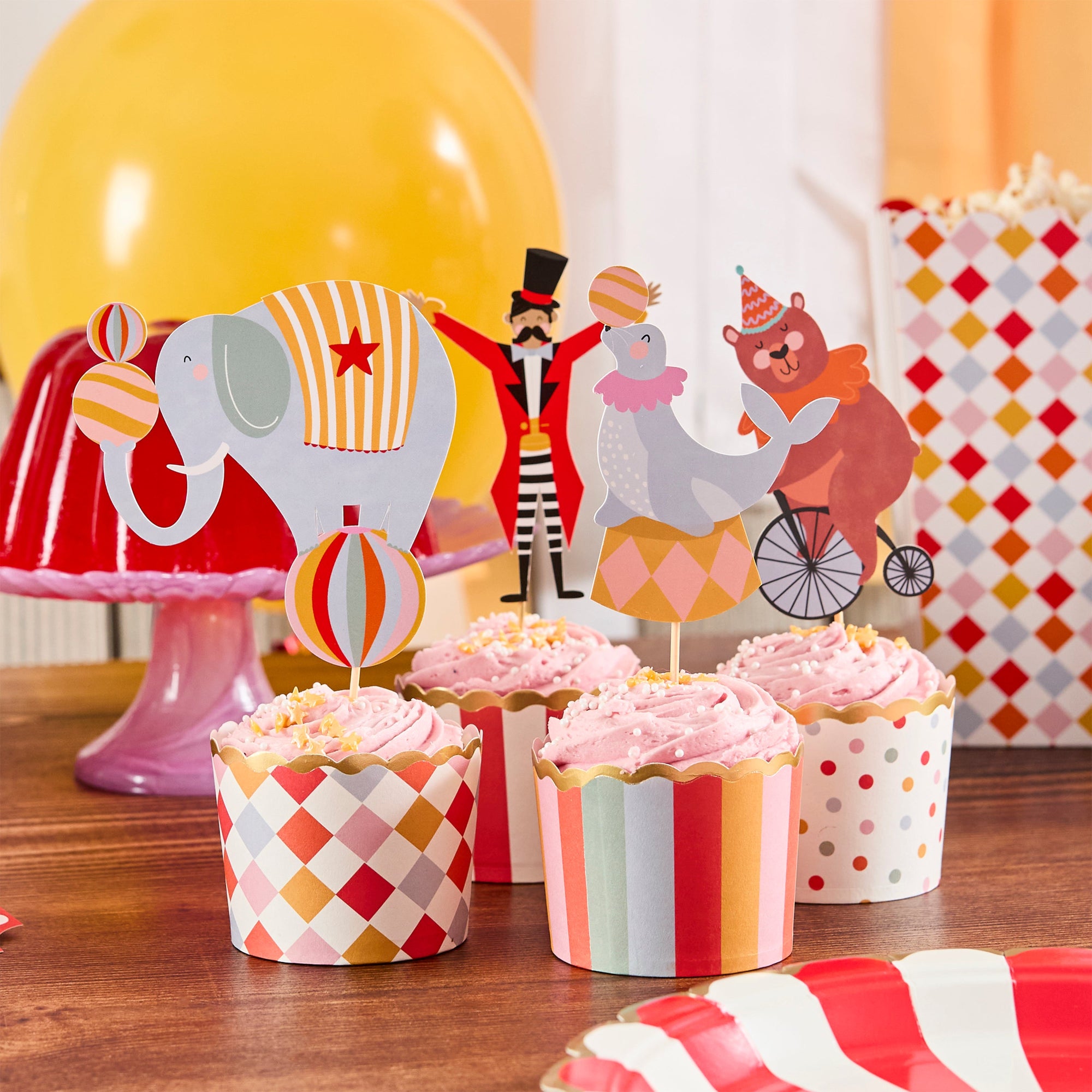 Four circus-themed treat cups with gold scalloped edges filled with pink-frosted cupcakes and topped with Ringmaster, elephant, and bear characters.