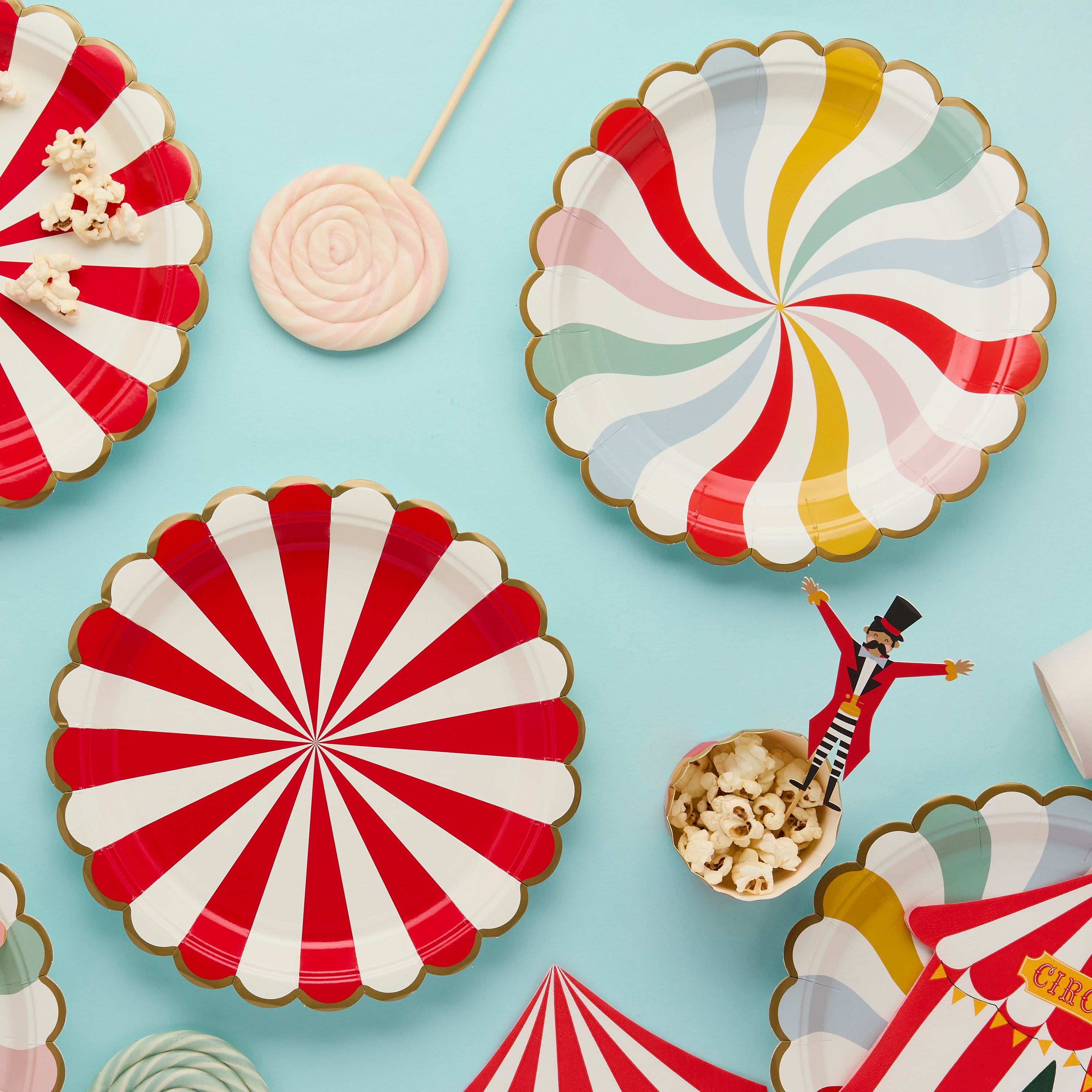 Circus paper plates at the center of a blue background, surrounded by lollipops, popcorn, and a ringmaster character decoration.