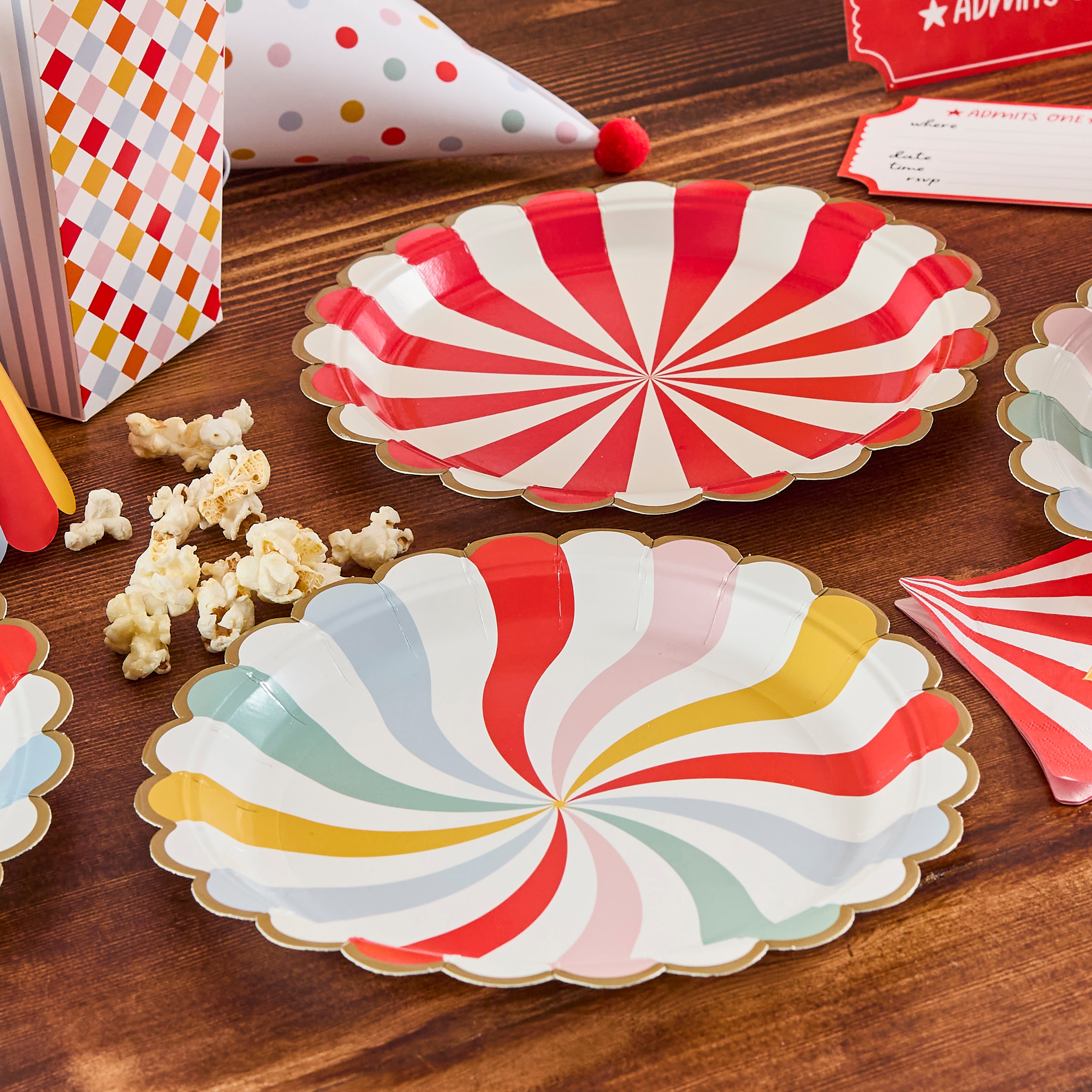 Vintage circus paper plates displayed on a wooden table next to popcorn boxes, party hats, and carnival invitations.
