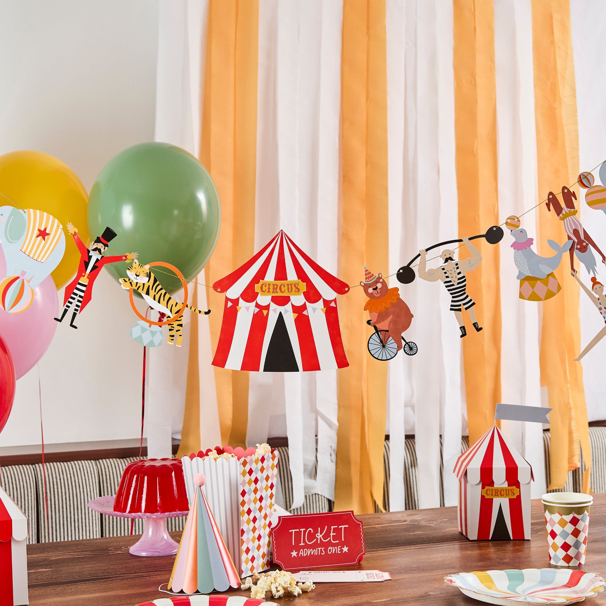 A full circus-themed party table set with popcorn, balloons, and a 3-meter vintage circus garland hung against a striped backdrop.