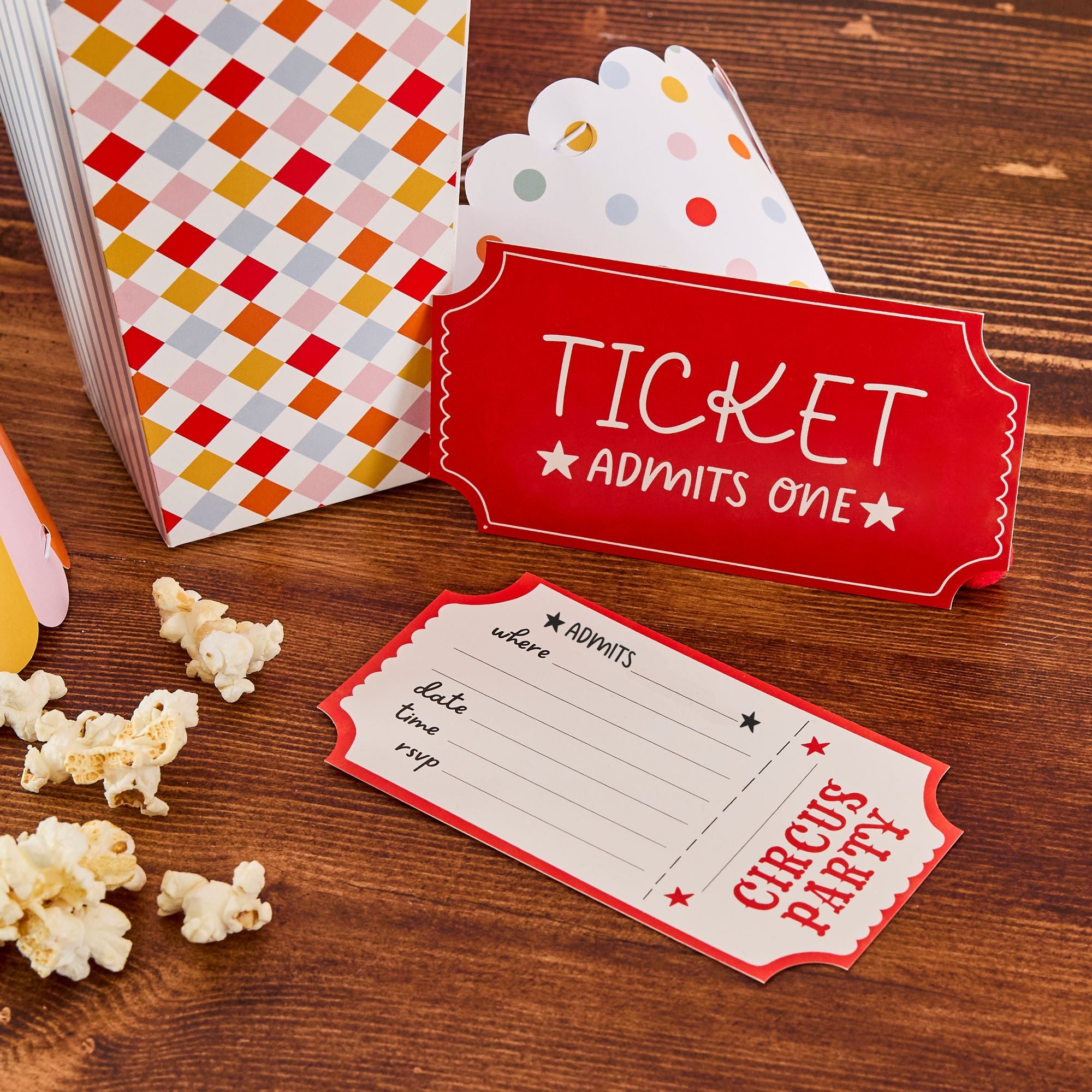 Big Top Setup: Circus ticket invitation sitting on a wooden table next to a diamond-patterned popcorn box and scattered popcorn.
