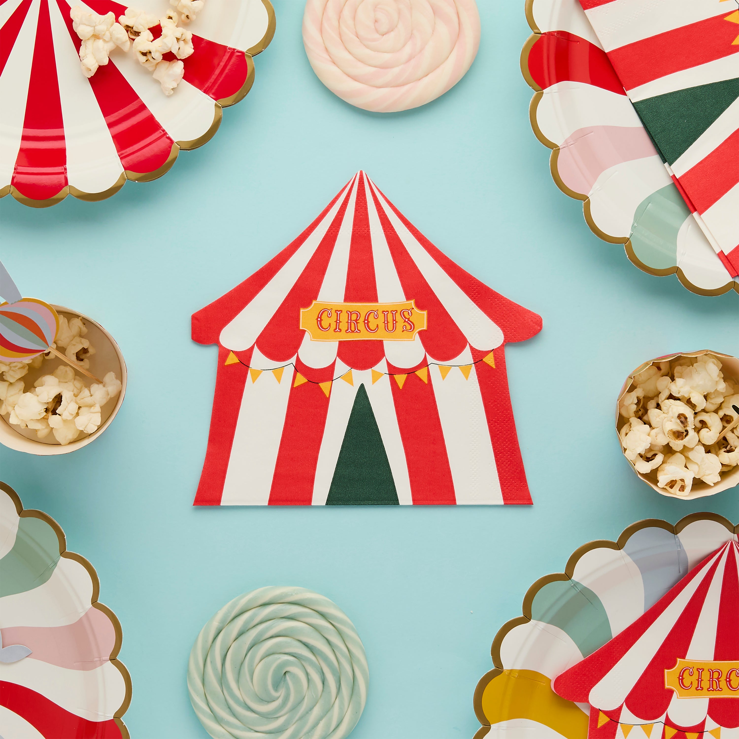A circus tent napkin at the center of a blue background, surrounded by lollipops, popcorn bowls, and scalloped carnival plates.