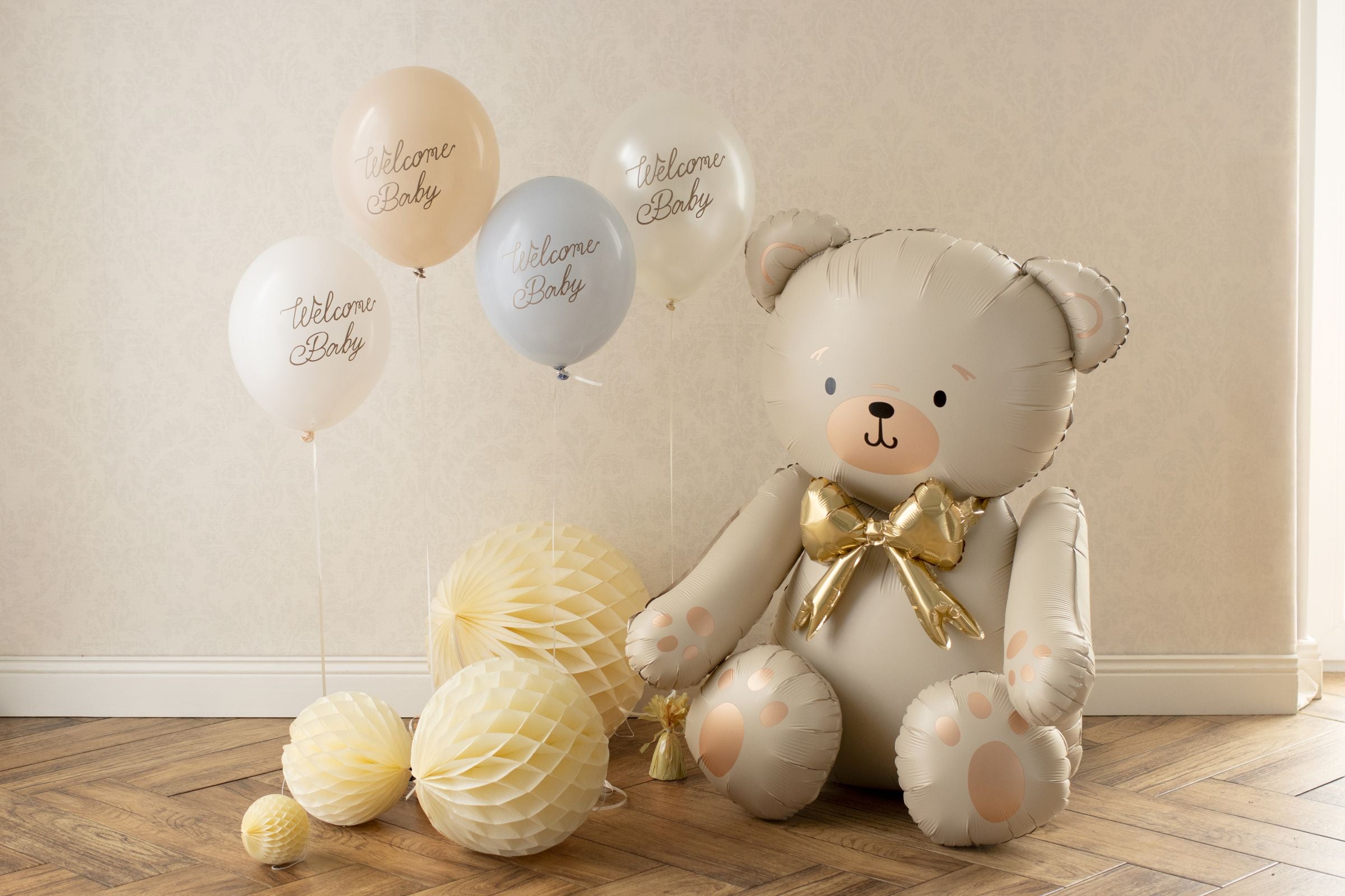 A large, cream-colored teddy bear foil balloon with a gold bow, sitting on a wooden floor next to a cluster of yellow honeycomb paper balls and a bouquet of pastel "Welcome Baby" balloons.