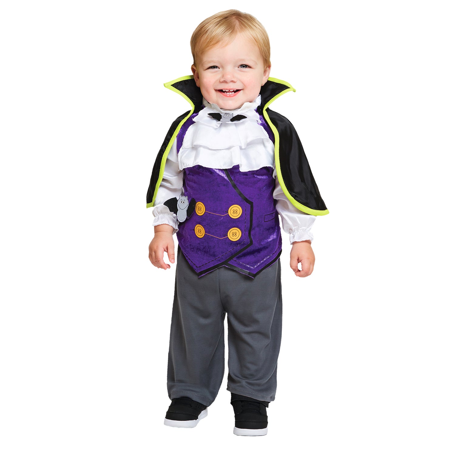 A smiling baby or toddler standing in a Dracula costume, which includes a black cape with a wide yellow and black collar, a white ruffled shirt, a purple vest with yellow buttons and a small ghost detail, and grey trousers.