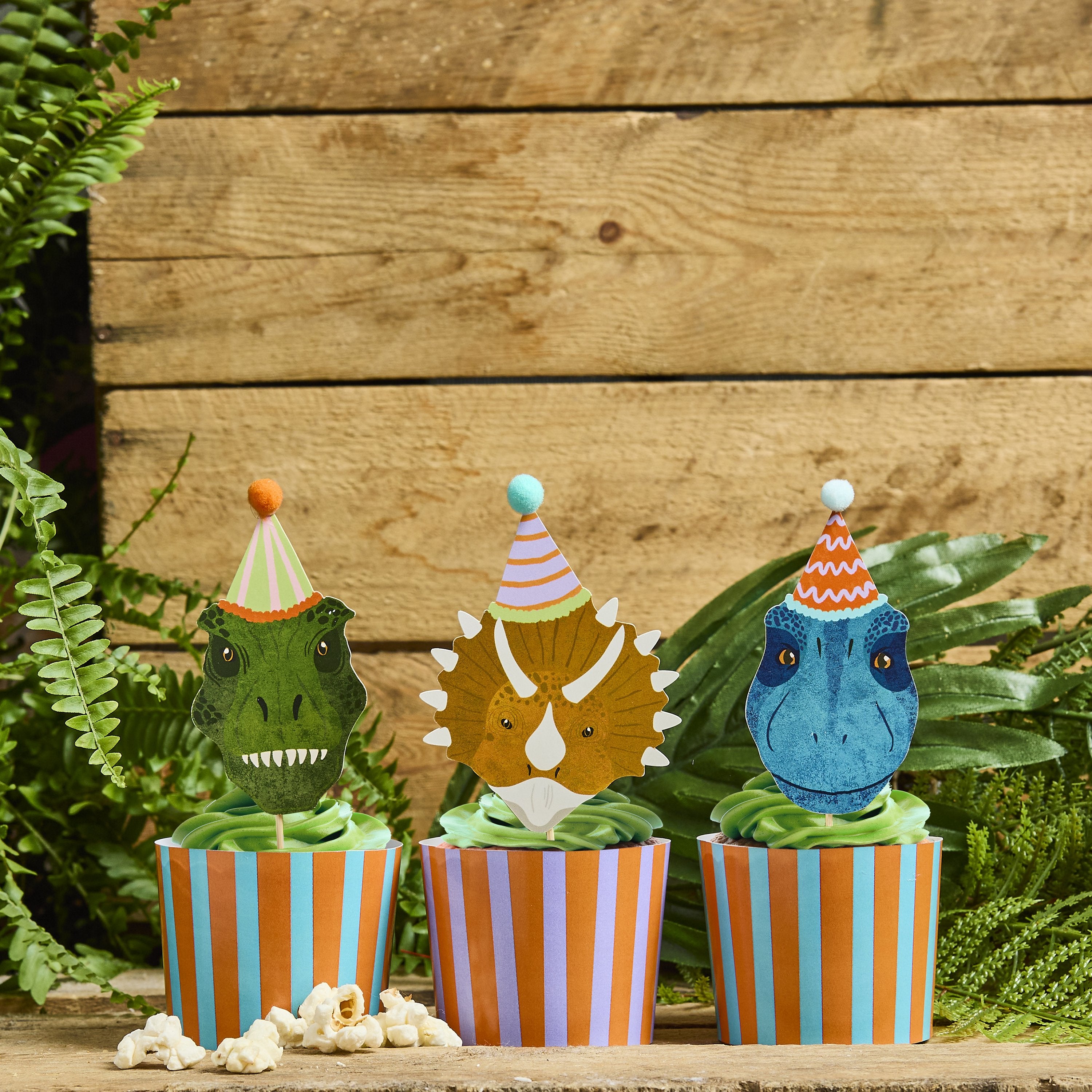 Three striped dinosaur party cups filled with green-frosted cupcakes and topped with T-Rex, Triceratops, and Raptor food picks with 3D pom-poms.