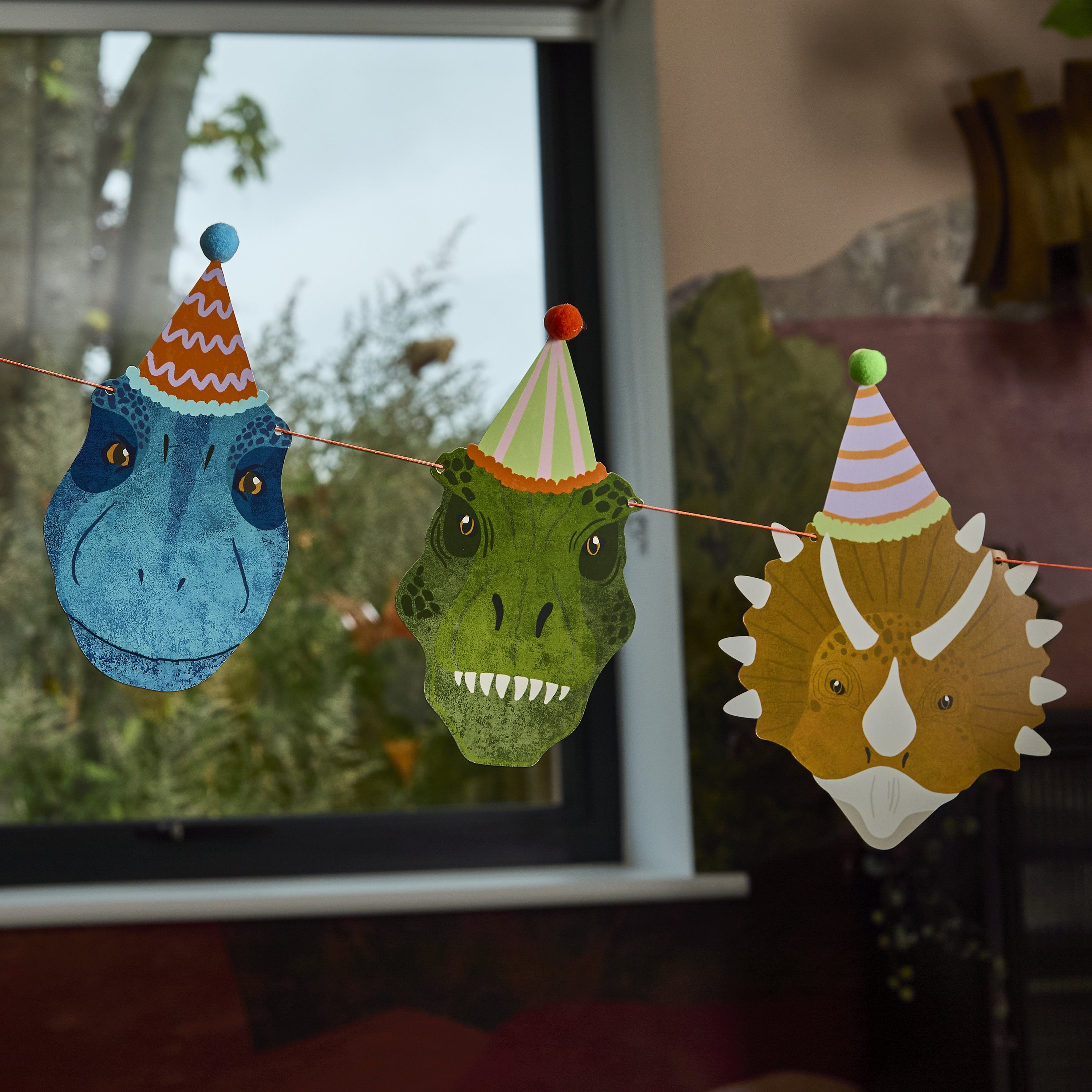 Close-up of dinosaur party bunting showing a blue T-Rex, green Raptor, and brown Triceratops with 3D pom-pom party hats.