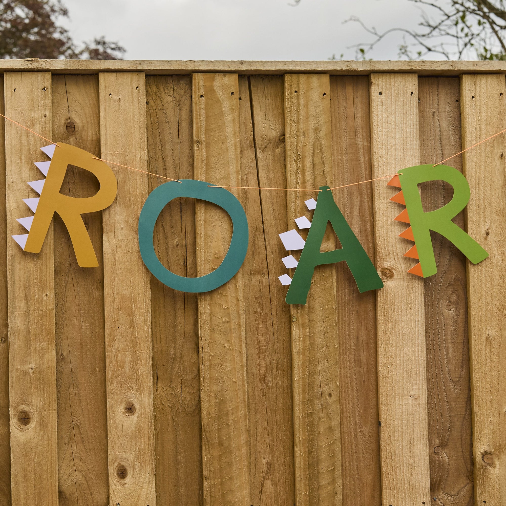 Dinosaur "ROAR" party banner hanging on a wooden garden fence, a legendary decoration for a prehistoric-themed outdoor birthday.