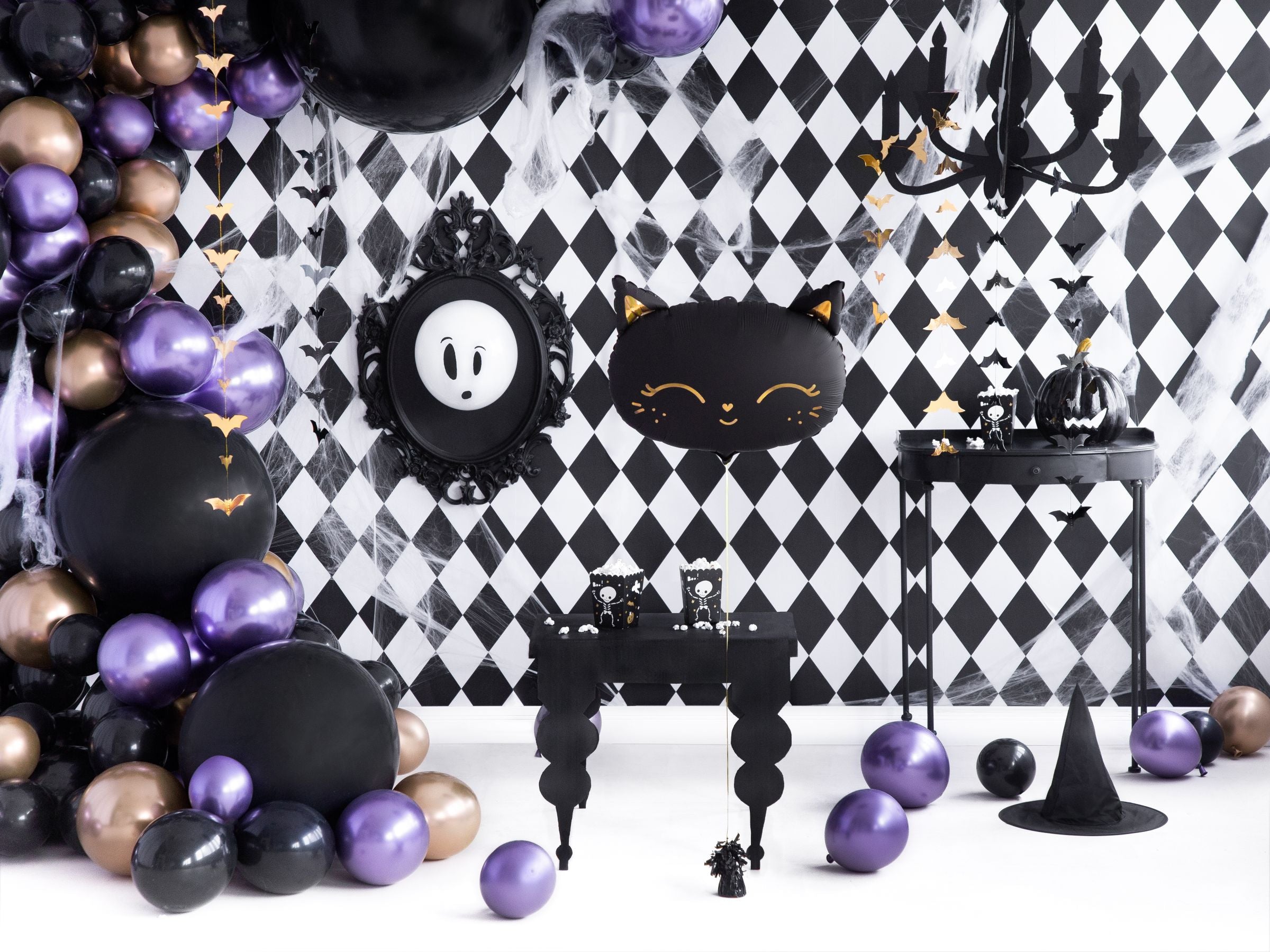 A stylish Halloween party backdrop with a black and white diamond pattern. The room is decorated with a black, purple, and gold balloon garland, a black table with treats, a black cat foil balloon with gold features, a framed ghost decoration, and a small black witch hat, with various balloons scattered on the floor.