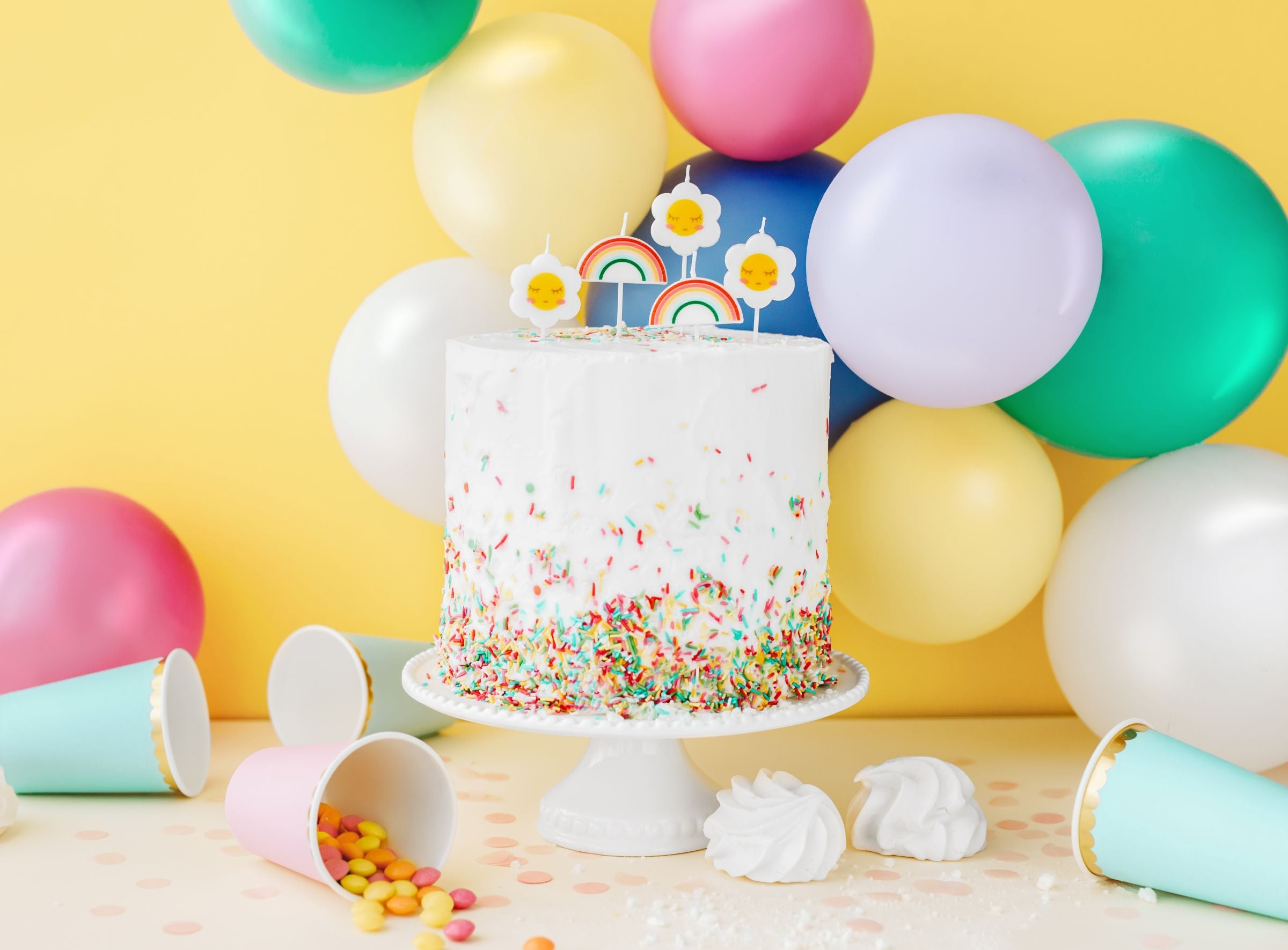 A birthday party scene with a white cake decorated with colorful sprinkles and rainbow candles. Various pastel balloons, including a fuchsia pink one, surround the cake against a yellow wall, along with party cups and other decorations.