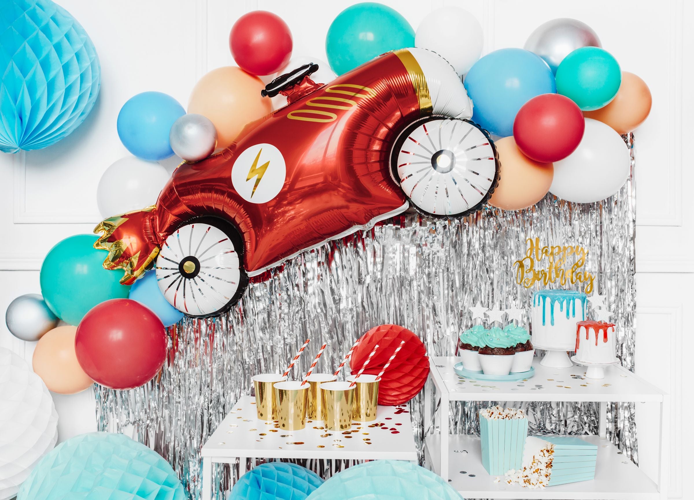 A colorful party setup with a red race car foil balloon mounted on a silver fringe backdrop. A garland of various balloons, including light blue, red, and orange, surrounds the car. On a table below, there's a birthday cake and party supplies like gold cups, popcorn boxes, and red striped straws.