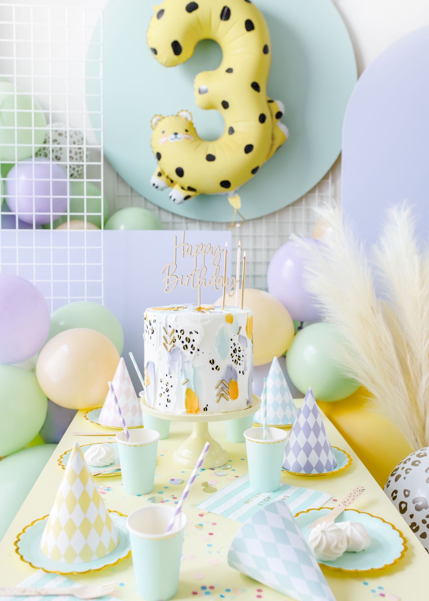 A close-up of a party table decorated for a third birthday celebration. The table features a modern birthday cake, pastel-colored cups and plates, small meringues, and is surrounded by a balloon arch of light peach, mint green, and lavender balloons. A large yellow number "3" foil balloon with leopard spots is visible in the background.
