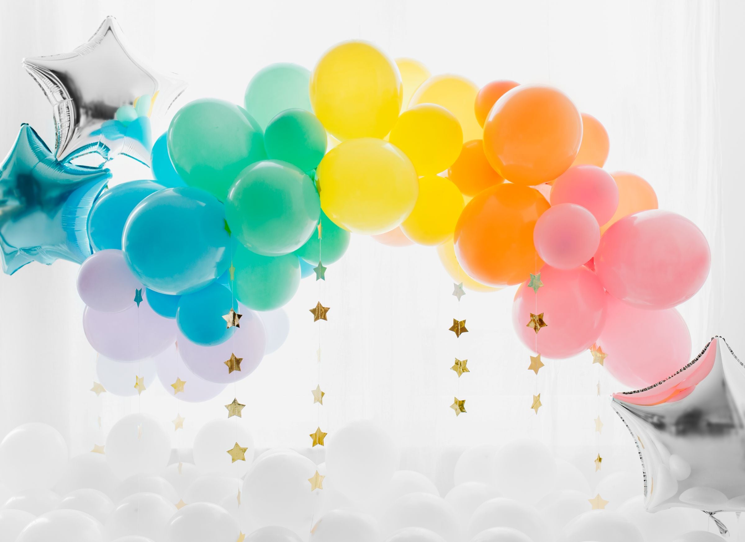 A vibrant balloon arch decoration in a rainbow of pastel colors, including pink, yellow, blue, and green, with hanging gold star confetti. The arch frames a backdrop of a white wall and is surrounded by a bed of white balloons on the floor, with silver star-shaped balloons at the sides.