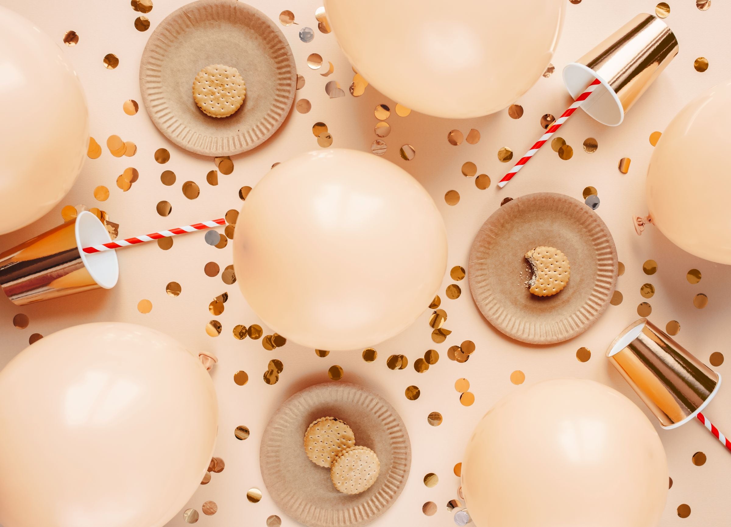 An overhead flat lay of a party scene with multiple inflated pastel peach balloons, paper plates with cookies on them, metallic gold cups with striped straws, and scattered gold confetti, all on a light peach background.