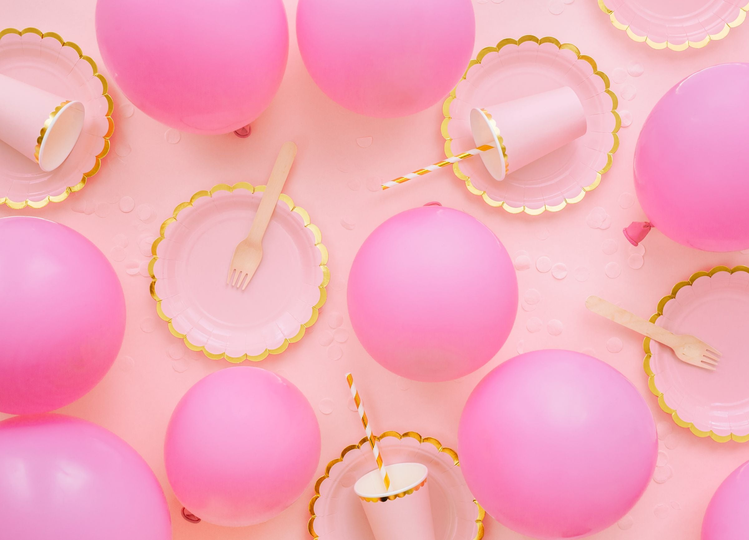 An overhead flat lay of a party scene with multiple pastel pink balloons, gold-rimmed pink scalloped plates, pink cups, wooden forks, and scattered confetti, all arranged on a pink background.