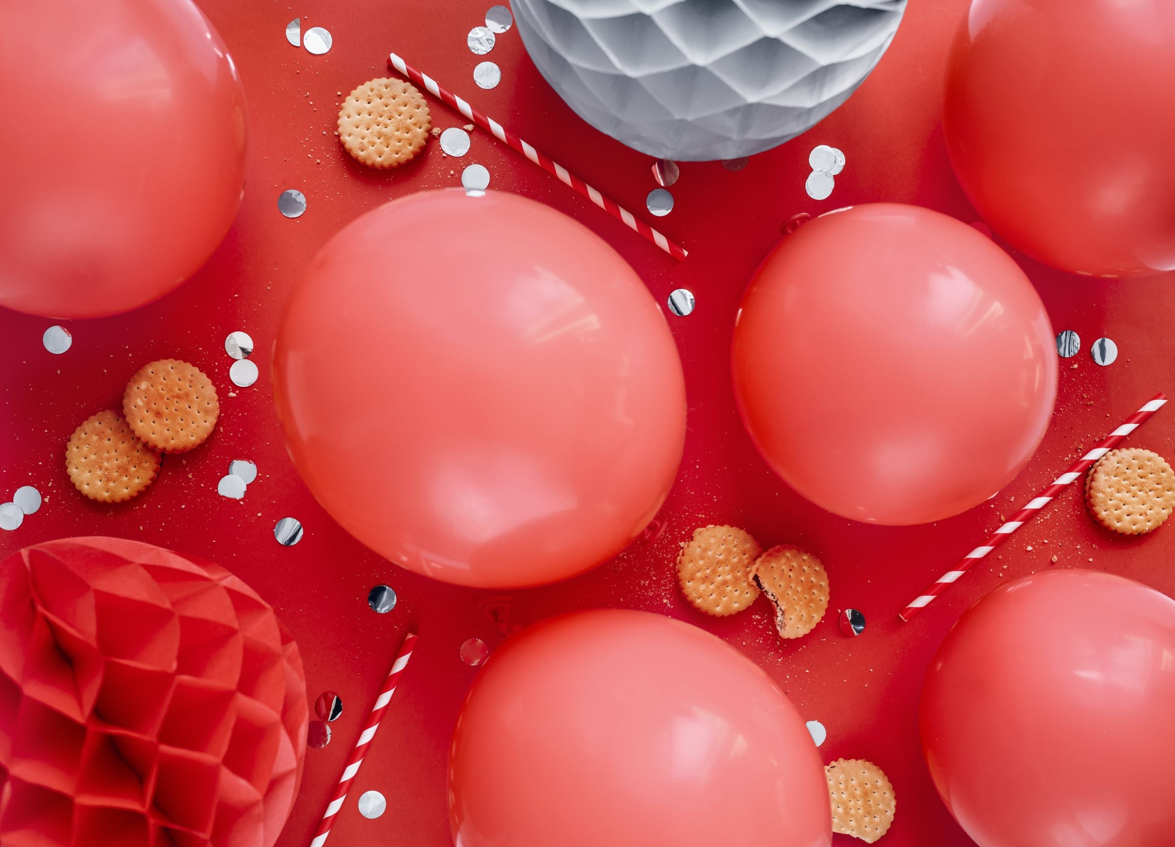 An overhead flat lay of a party scene on a red background, featuring multiple inflated pastel red balloons, red and white striped straws, small round cookies, and honeycomb paper balls, along with scattered confetti.