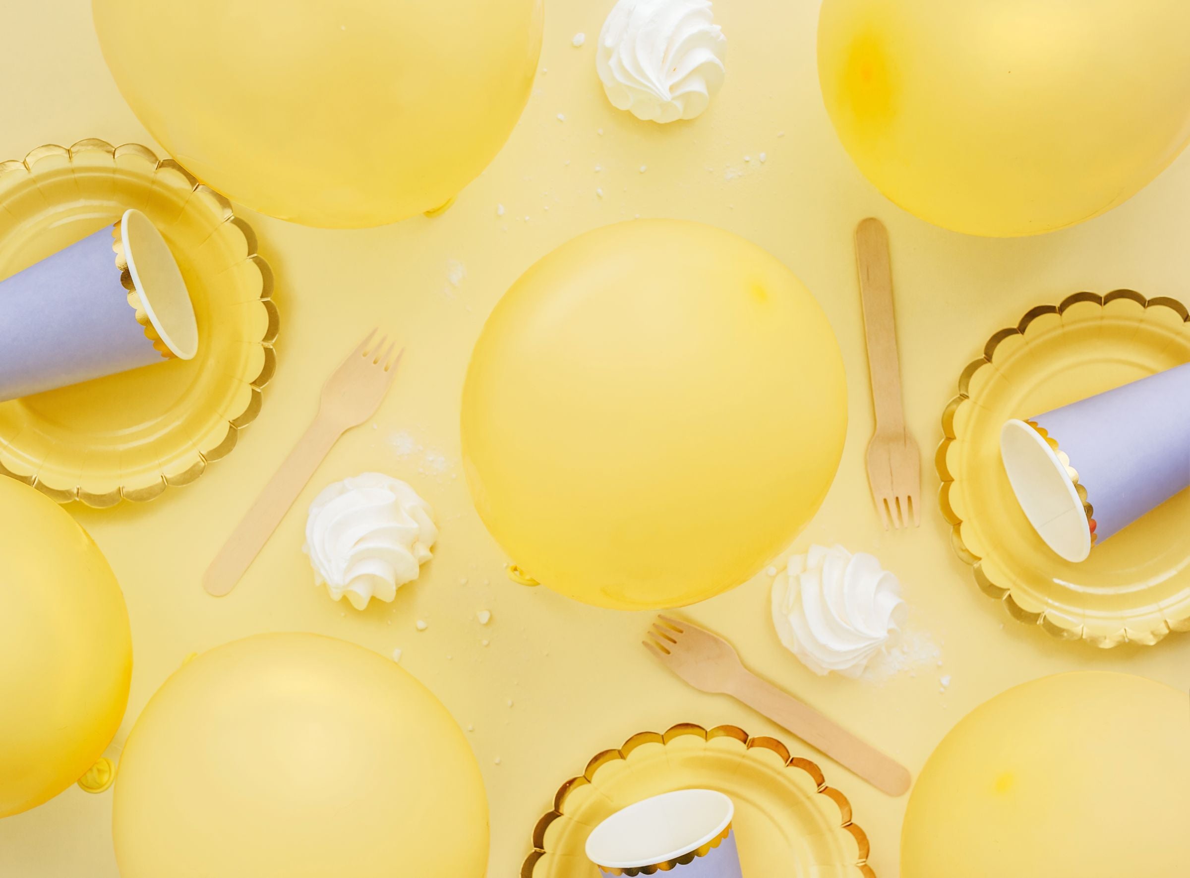 An overhead flat lay of a party scene with pastel yellow balloons, gold-rimmed plates, wooden forks, and white meringues, all arranged on a yellow background with scattered white powder.