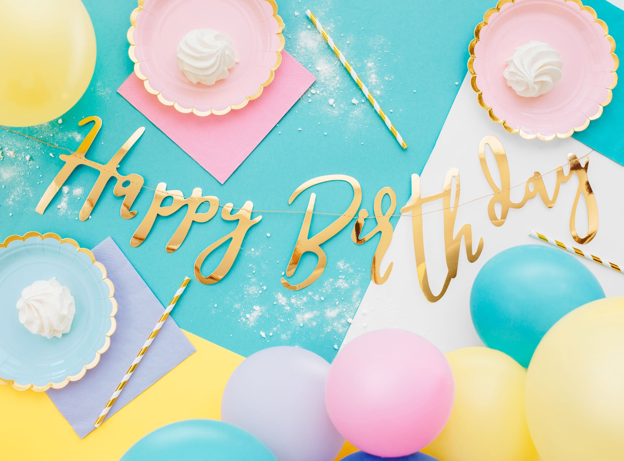 A close-up of a birthday party table featuring a white cake with colorful sprinkles and rainbow-themed candles, surrounded by various pastel-colored balloons, including yellow, pink, blue, and mint green.