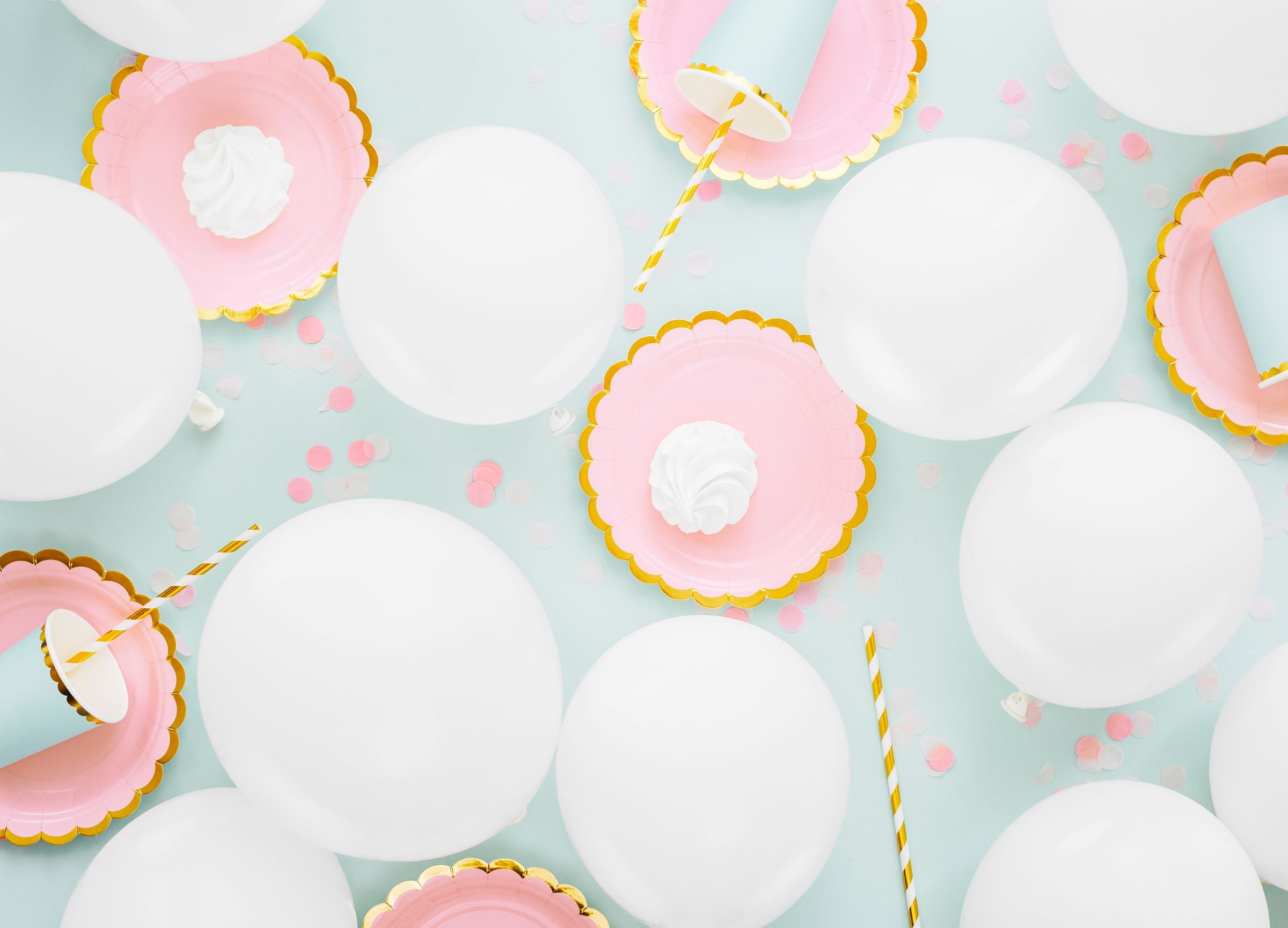 An overhead flat lay of a party scene on a light blue background, featuring multiple inflated white balloons, pink scalloped plates with meringues, and light blue cups with gold striped straws, along with scattered confetti.