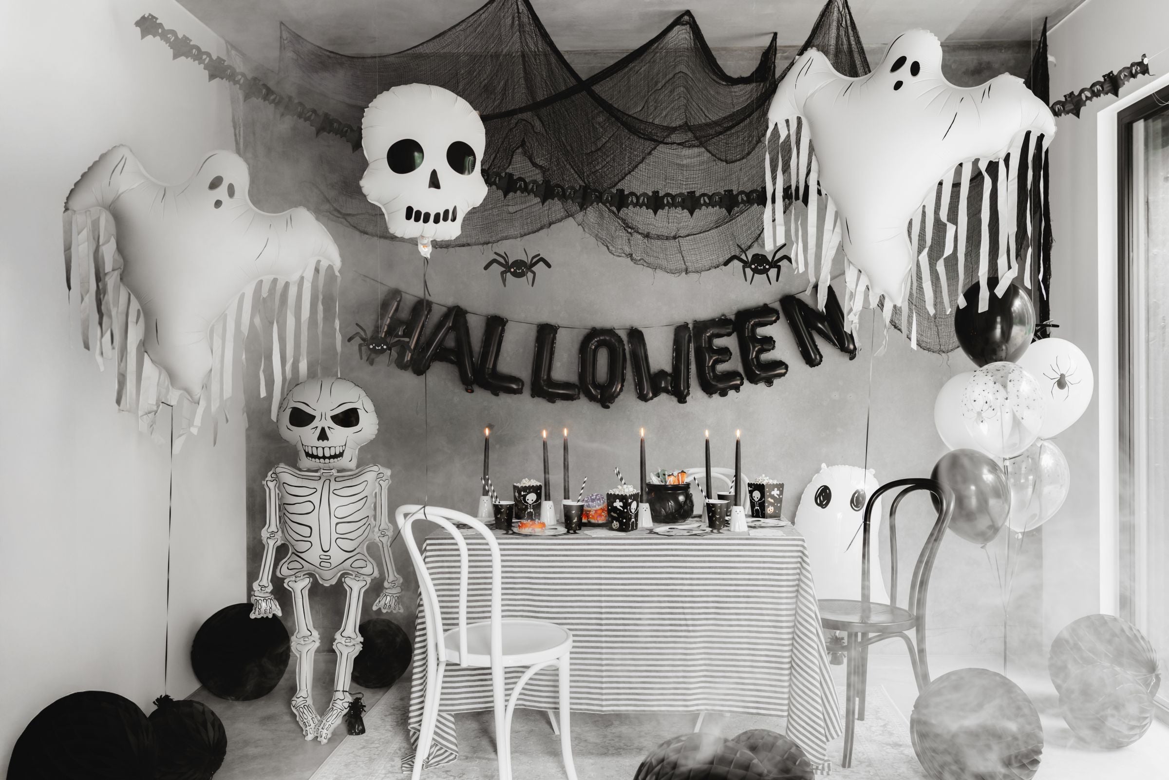 A Halloween party scene with a table set with themed tableware, candles, and chairs. In the background, black netting with spiders, a skull balloon, a "HALLOWEEN" letter balloon banner, and multiple white foil ghost balloons with tassels are visible, creating a spooky atmosphere.