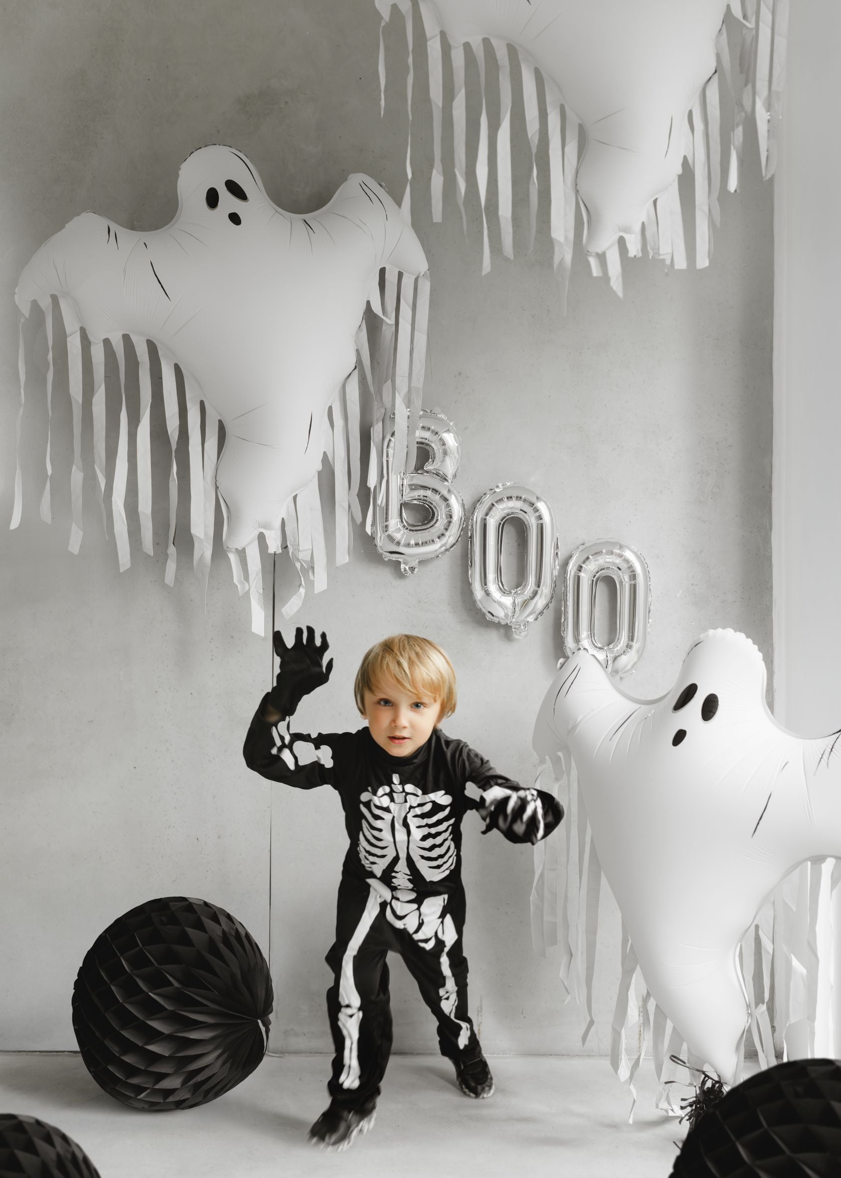 A child dressed in a black skeleton costume, posing with hands raised, in a Halloween-decorated room. Large white foil ghost balloons with tassels hang from the ceiling, along with silver "BOO" letter balloons and black honeycomb balls.