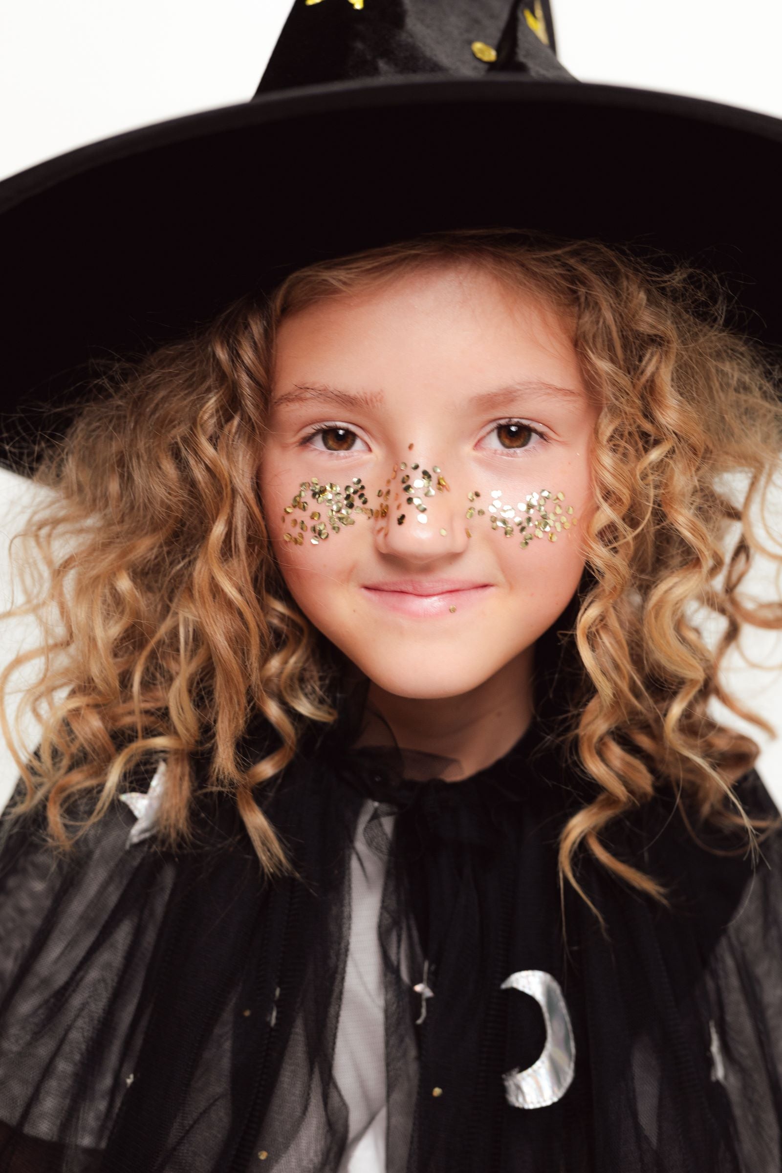 A close-up of a child's face, smiling, with gold glitter applied across the bridge of the nose and cheeks to create a freckle effect, and wearing a black witch hat.