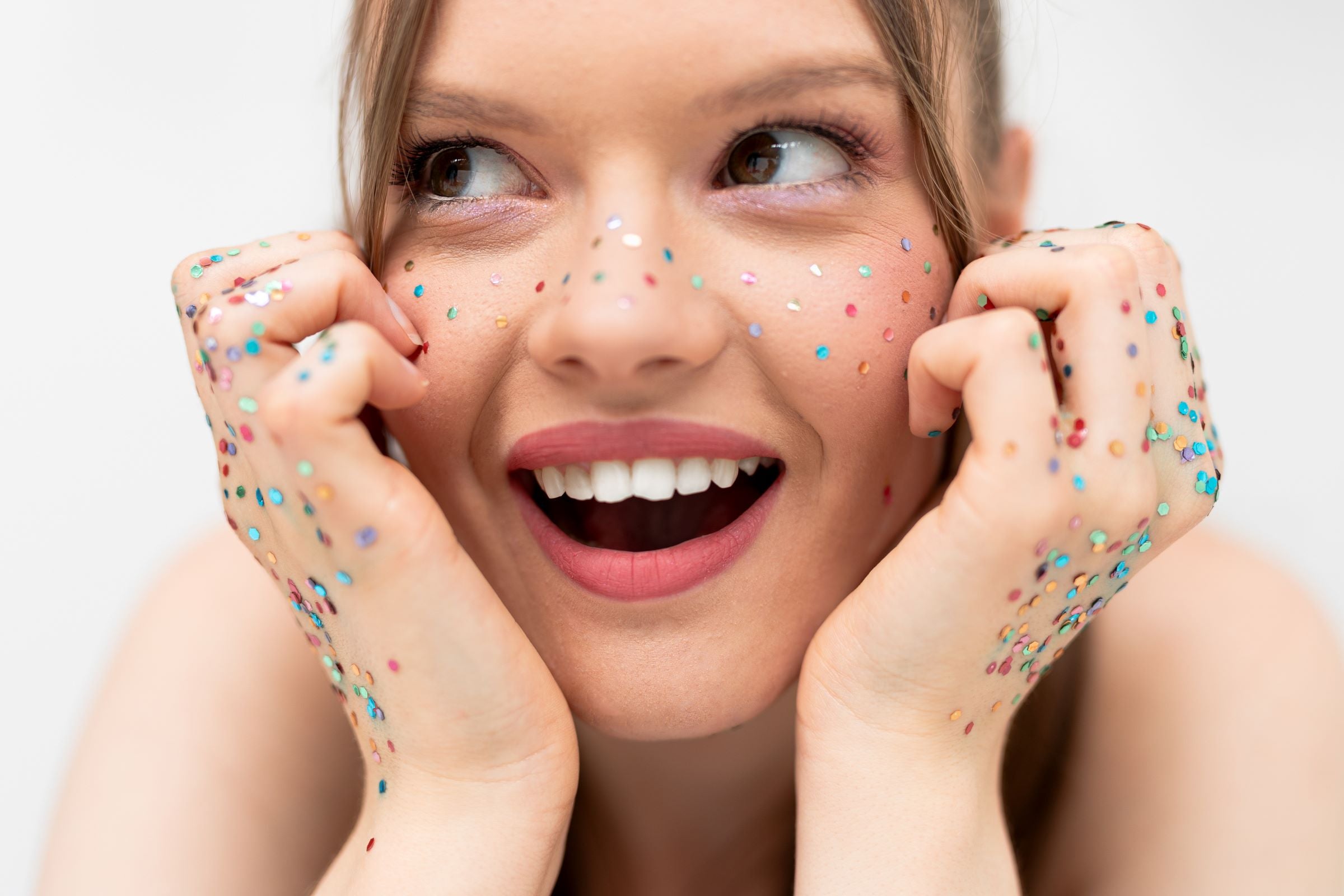 A close-up of a person's face smiling, with multicoloured glitter applied in a freckle pattern across the nose and cheeks. The person's hands are also covered in glitter and are held up near their face.