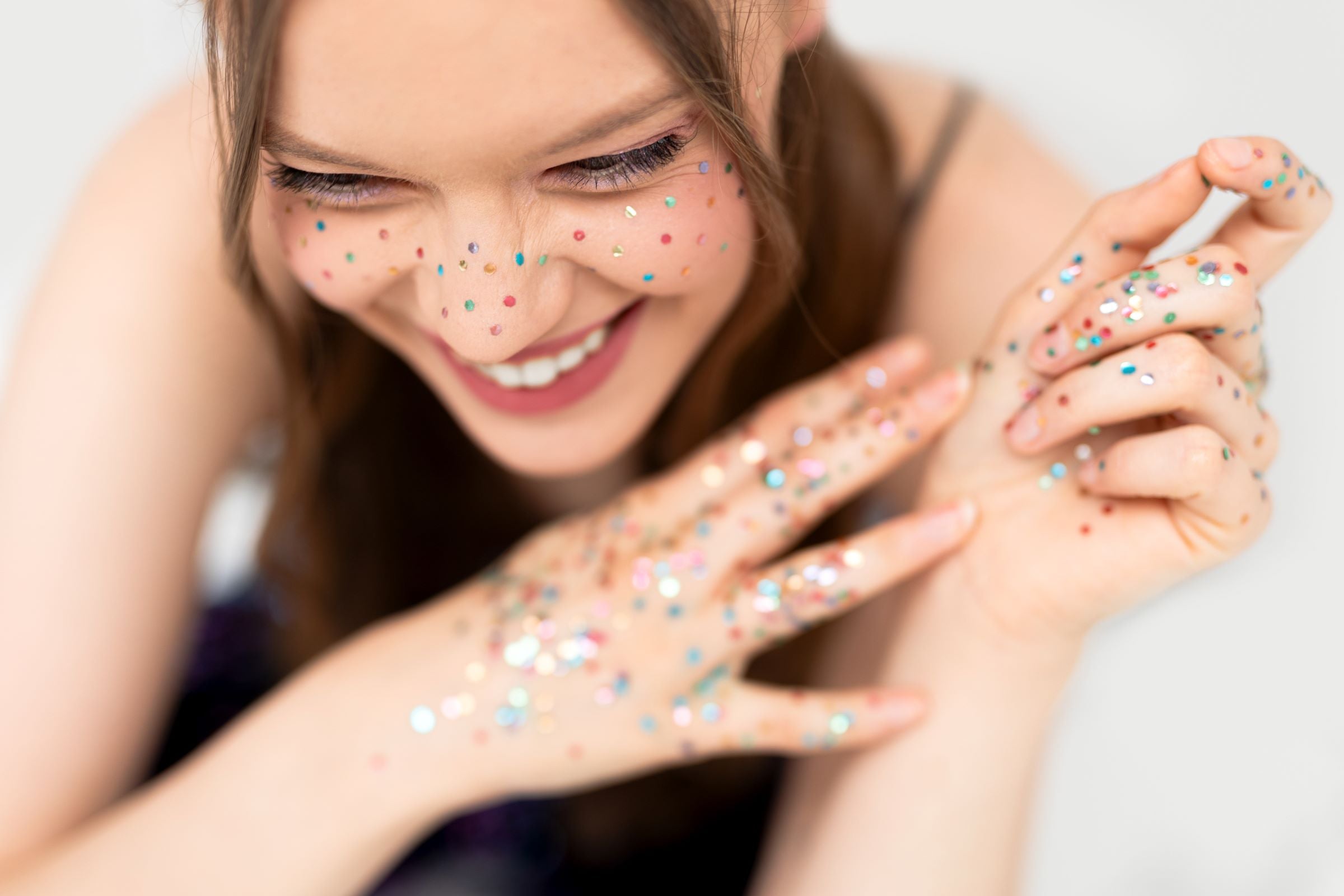 A close-up of a person's face with their eyes closed and smiling, with multicoloured glitter applied in a freckle pattern. The person's hands, also covered in glitter, are visible in the foreground.