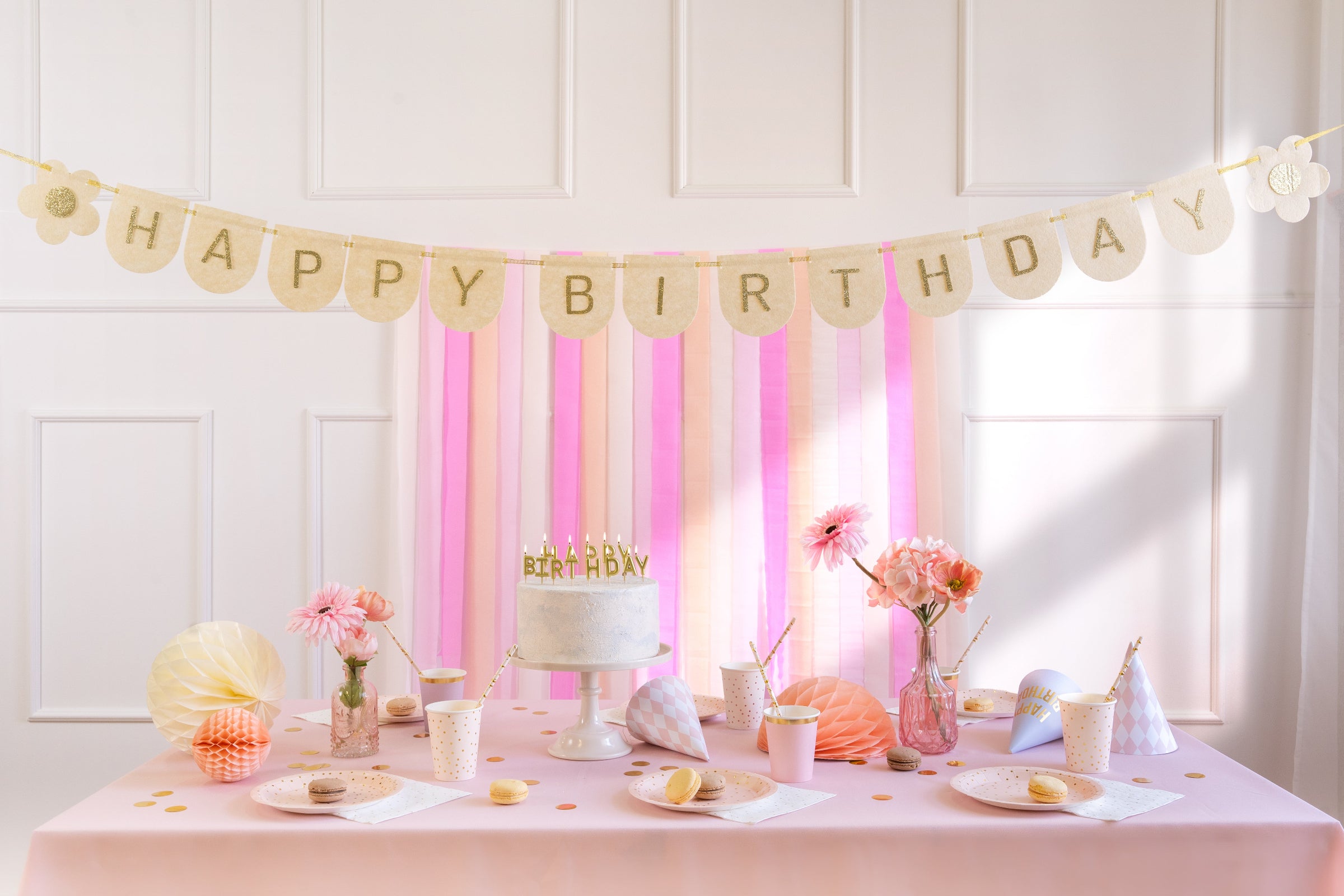 A beautifully decorated birthday party table with pink accents, featuring a white cake, fresh flowers, and various partyware, with the "HAPPY BIRTHDAY" felt banner with glitter gold letters hanging above it against a light-colored wall with pink and white striped backdrop.
