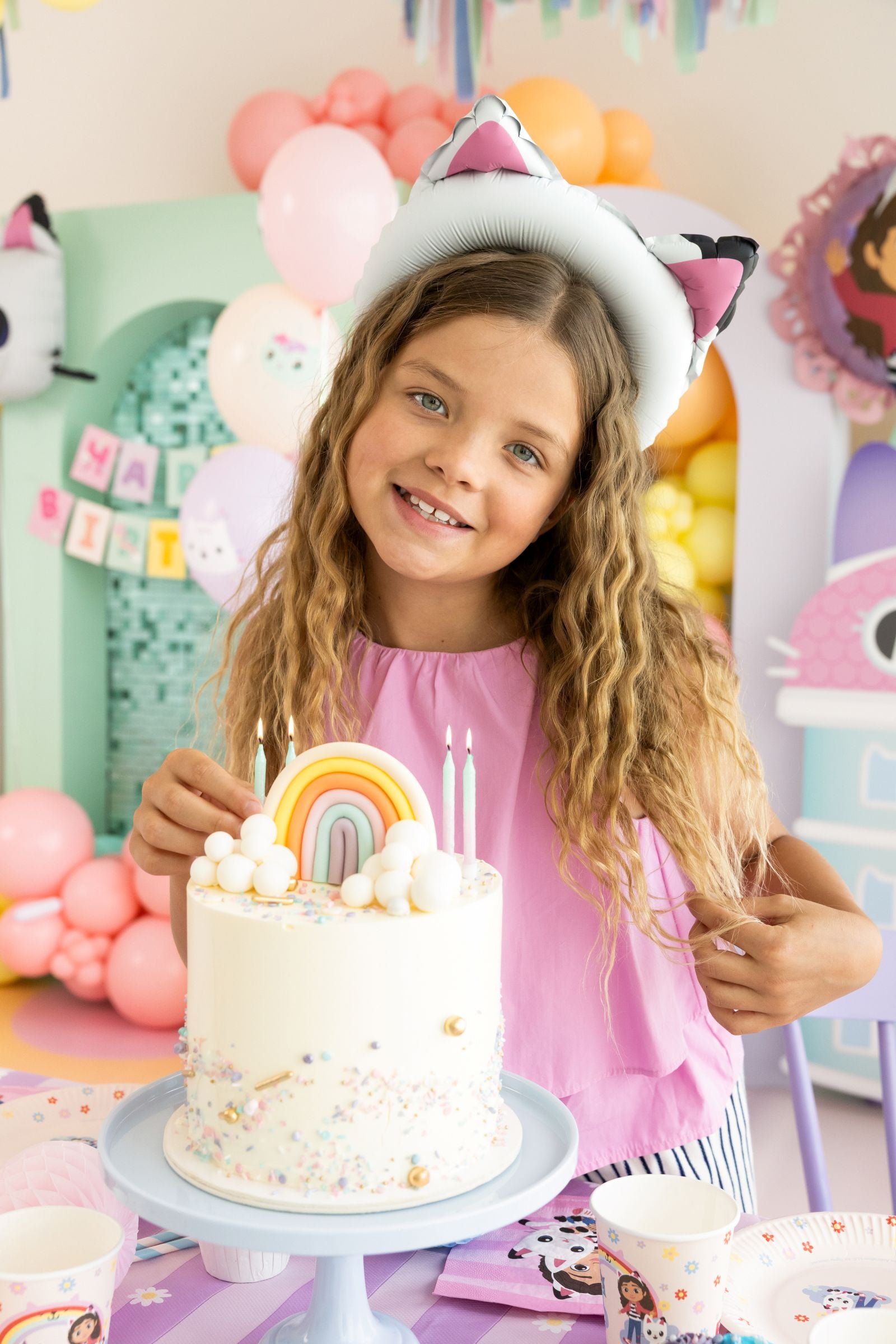 A child wearing cat ear balloons sitting next to a rainbow cake in a fully decorated Gabby's Dollhouse themed room.