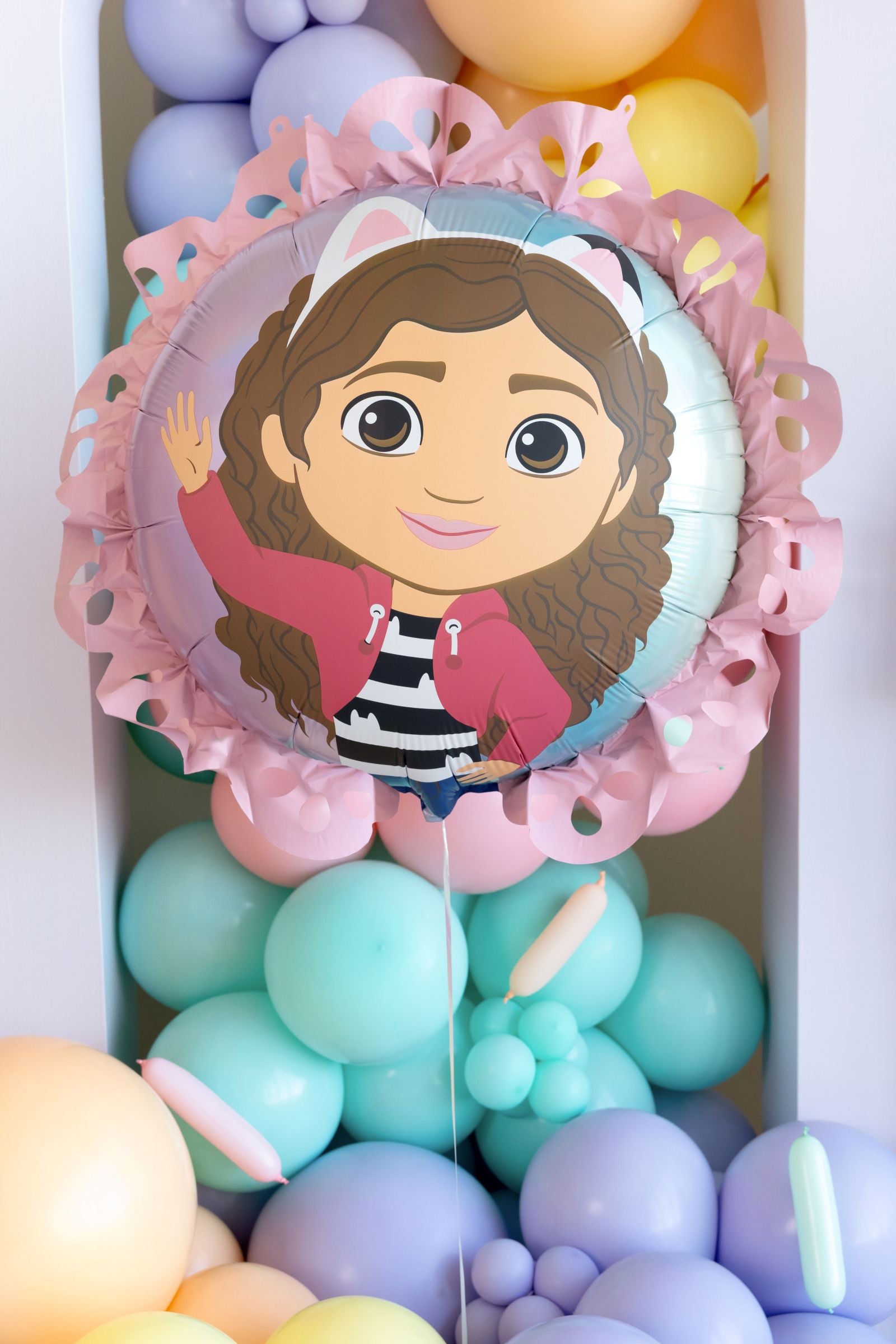 Round Gabby’s Dollhouse foil balloon featuring Gabby with a decorative pink paper ruffle edge surrounded by colorful balloons