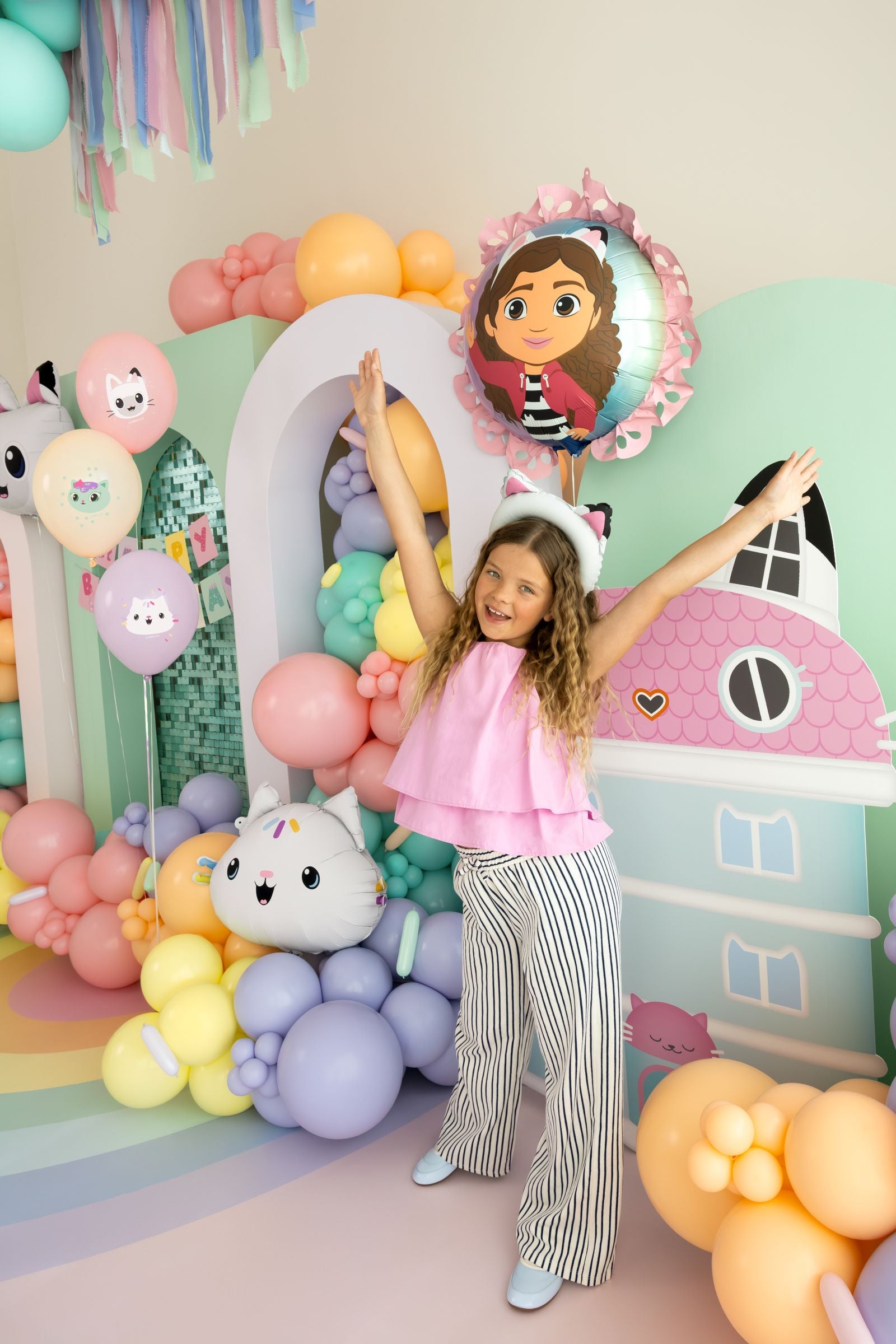 A young girl posing happily next to a ruffled Gabby's Dollhouse character balloon in a festive party room.