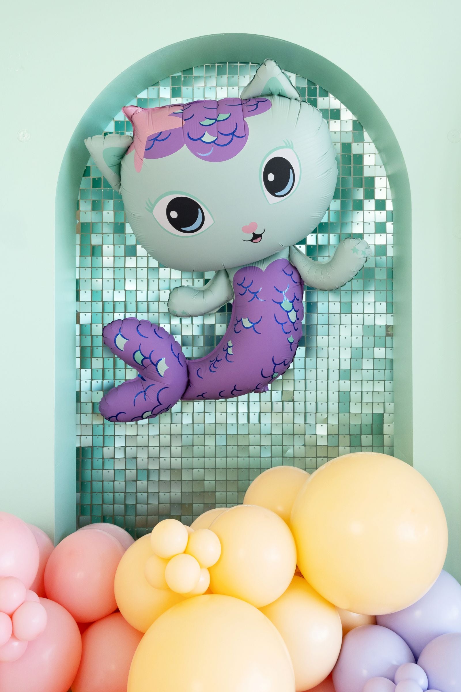 Giant MerCat shaped foil balloon with teal and purple mermaid scales and a starfish on head against a light green mosaic wall with colorful balloons.