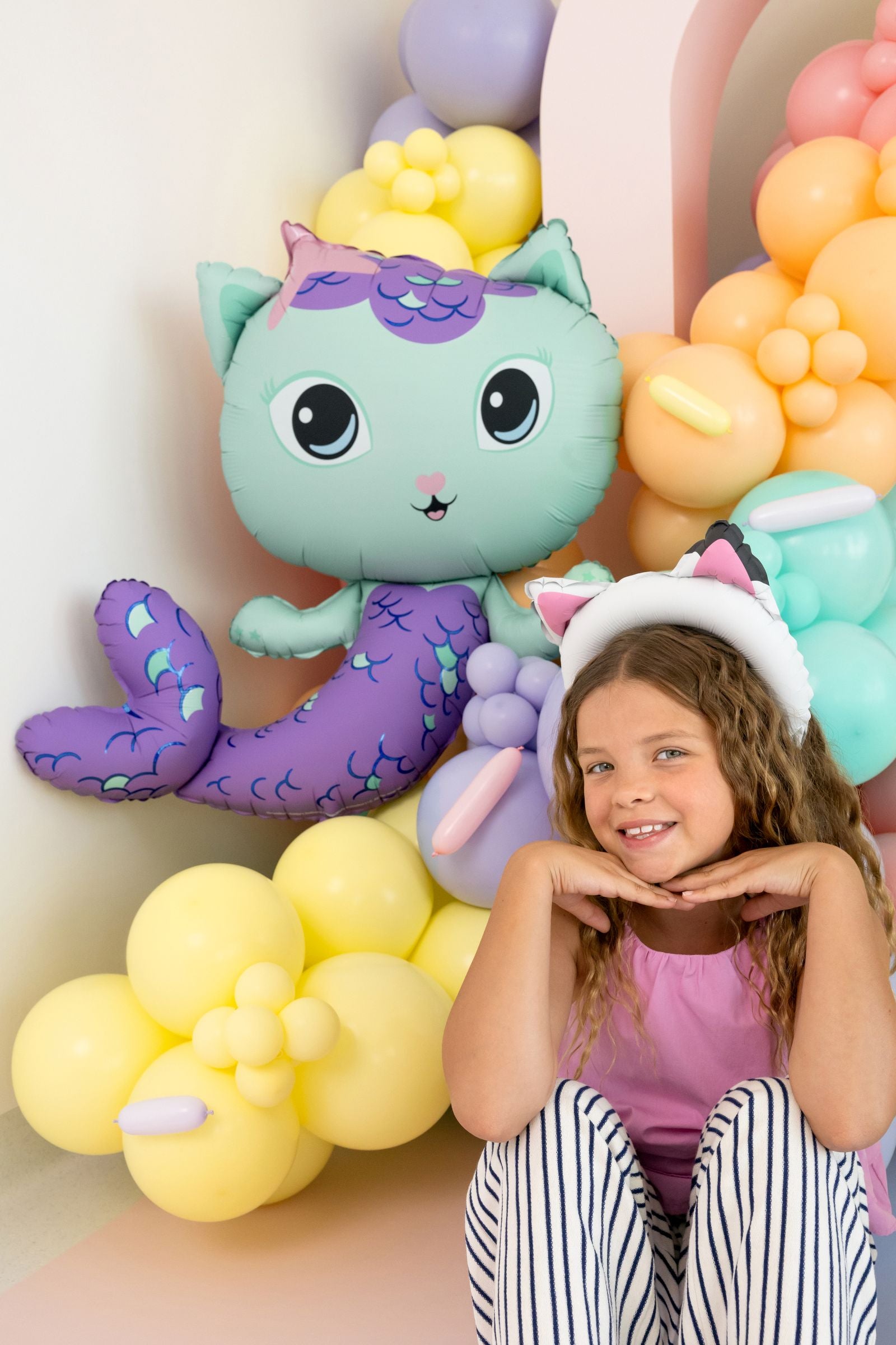 A happy child sitting next to a large MerCat foil balloon at a Gabby's Dollhouse themed party.