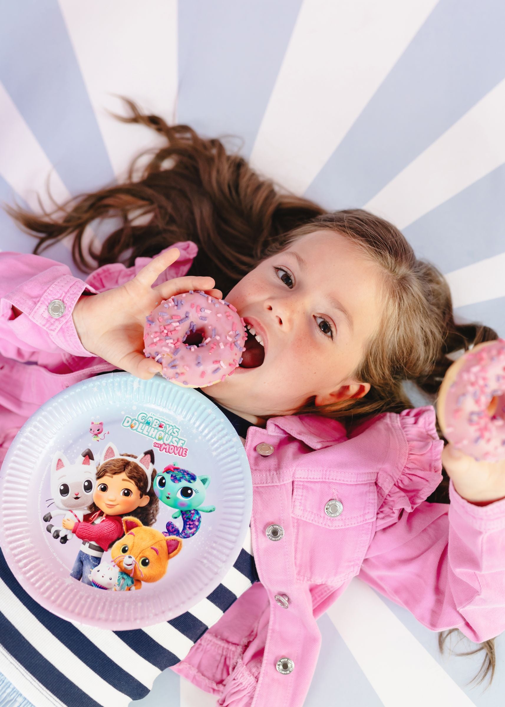 A smiling child in a pink jacket lying on a striped surface, playfully holding two pink frosted donuts near their face alongside a Gabby's Dollhouse The Movie paper plate.