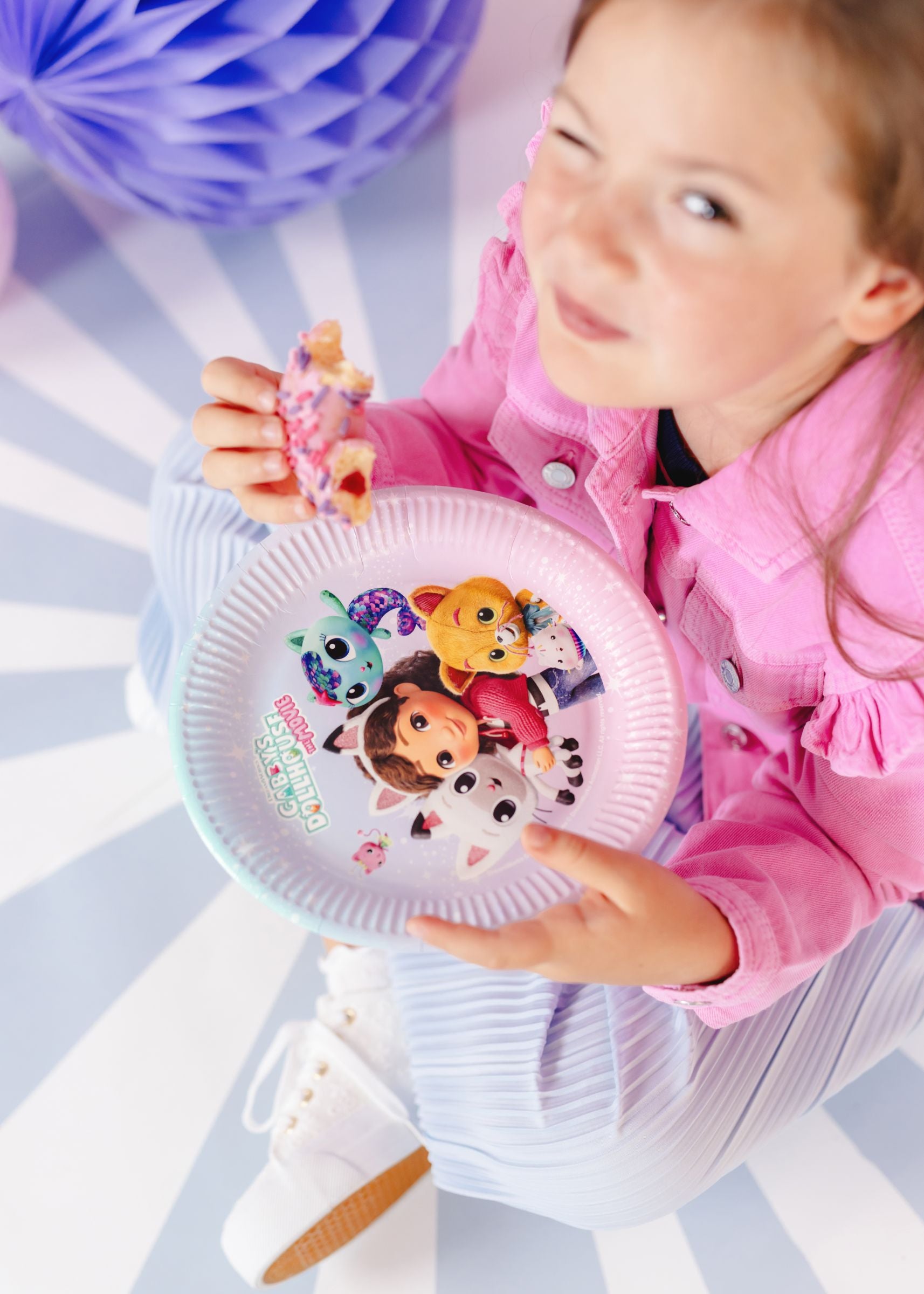 A child wearing a pink denim jacket sitting on a blue and white striped floor, holding a pink frosted donut and a Gabby's Dollhouse The Movie paper party plate.