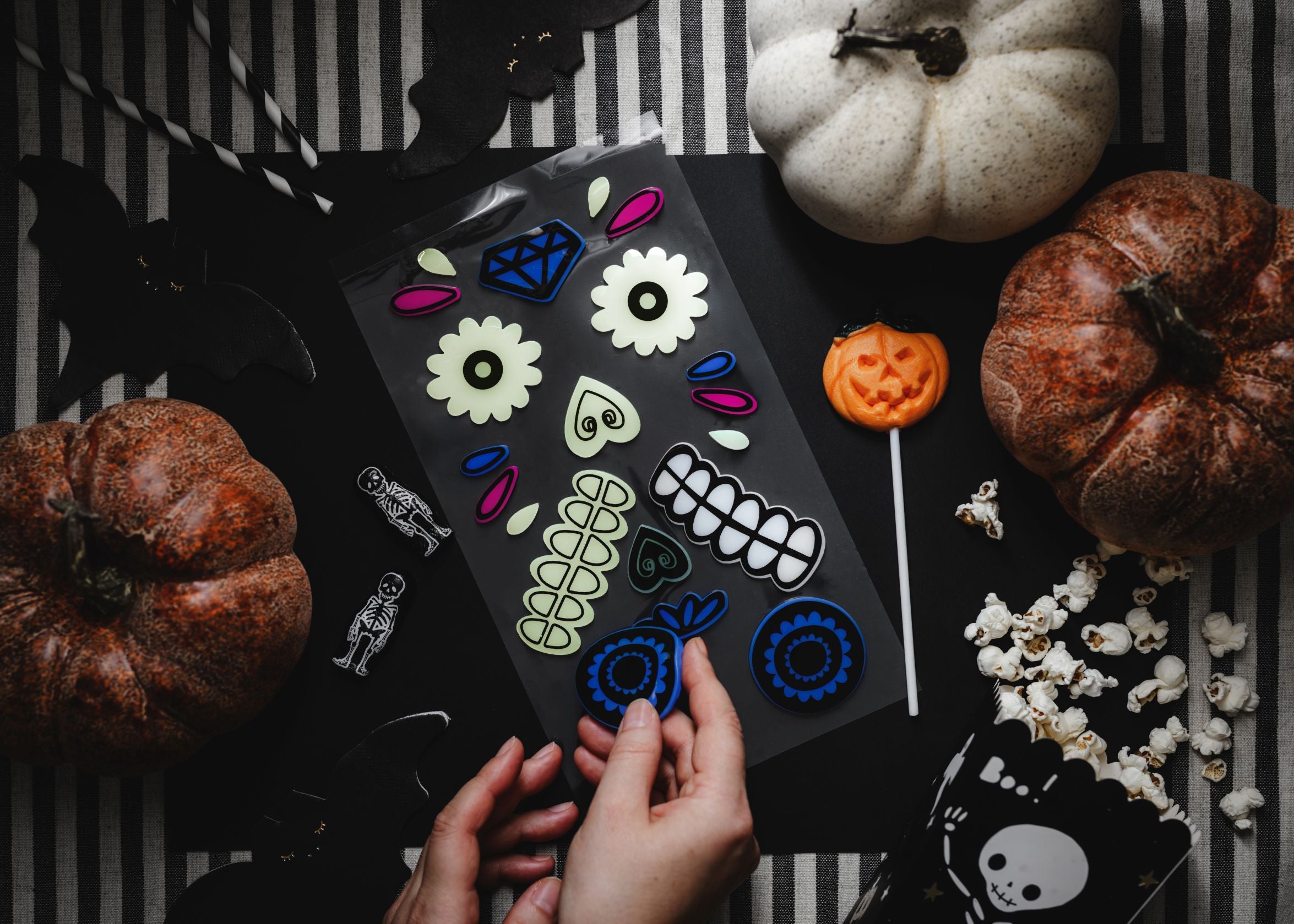 Hands are seen peeling a glow-in-the-dark gel sticker from a sheet to decorate an orange pumpkin, set on a black and white striped tablecloth with other Halloween and Day of the Dead themed decorations like black paper bats, small pumpkins, and popcorn in a skull-themed box.