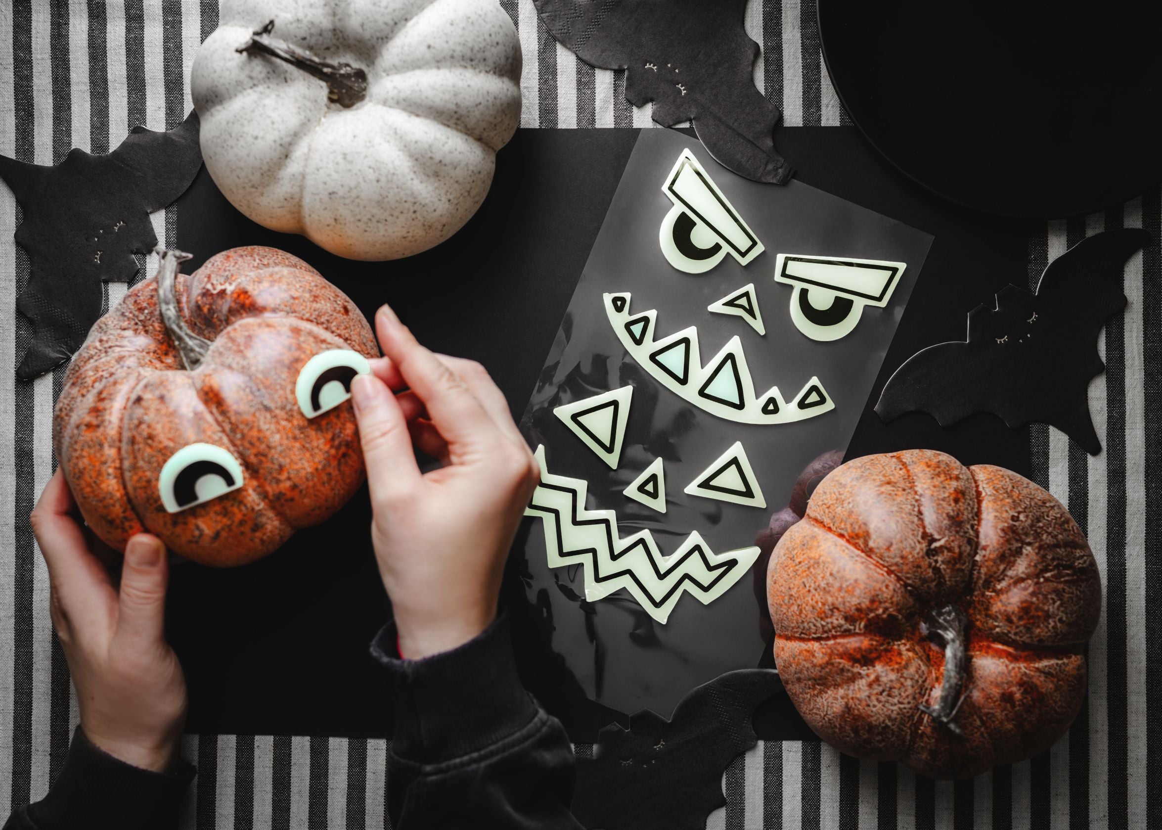 Hands applying a black and green-yellow gel sticker eye to a small orange pumpkin, set on a black and white striped tablecloth. Another unapplied sheet of matching gel pumpkin face stickers and two decorative pumpkins (one white, one orange) are also visible on the table.