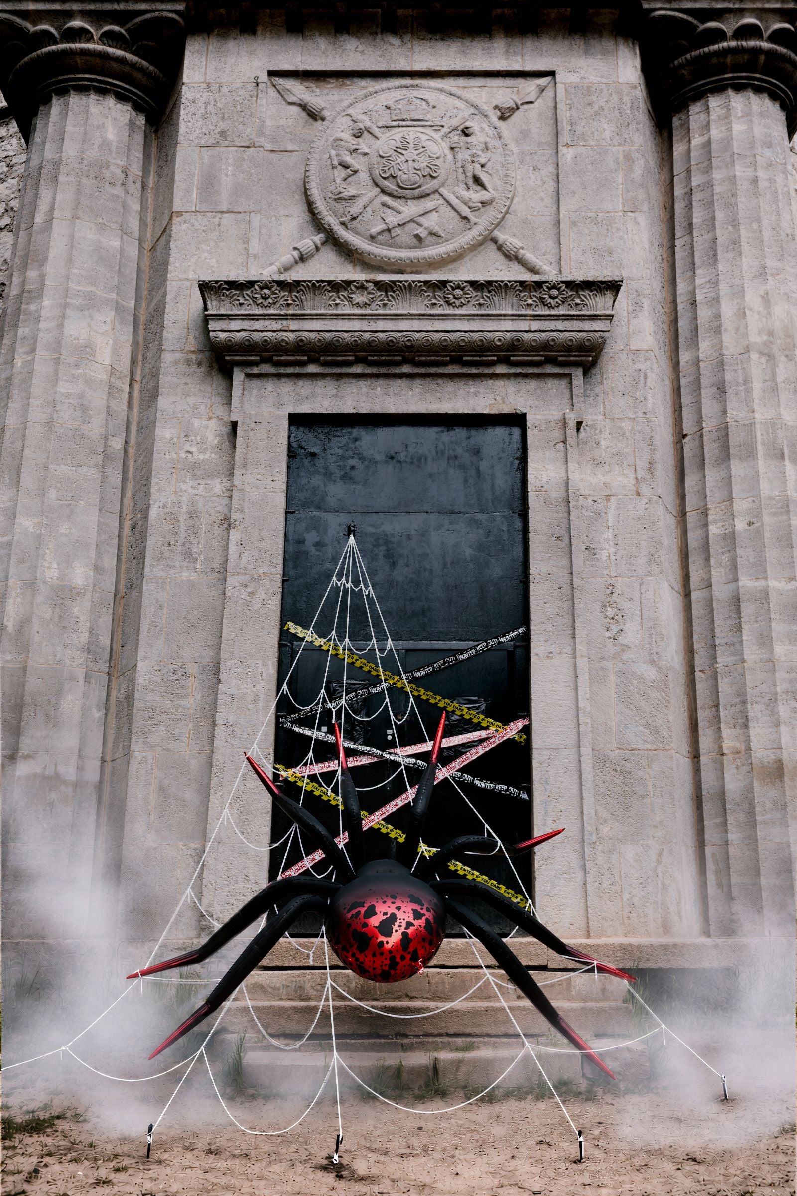 Giant standing spider foil balloon placed outdoors in front of a stone doorway covered in a white web and caution tape.