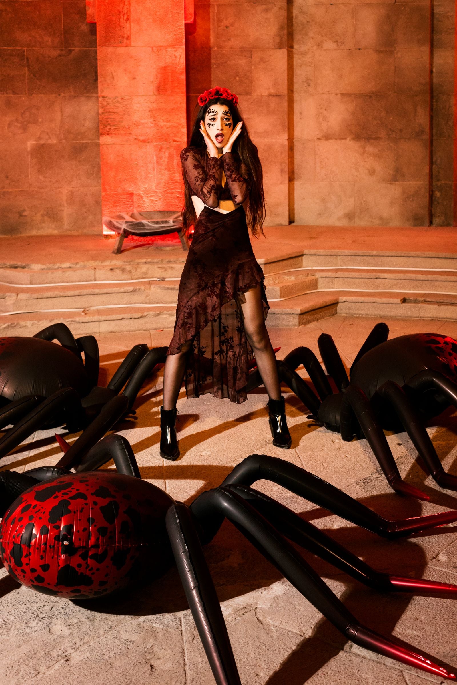 Woman in a dark dress standing looking shocked, surrounded by multiple giant black and red standing spider foil balloons.