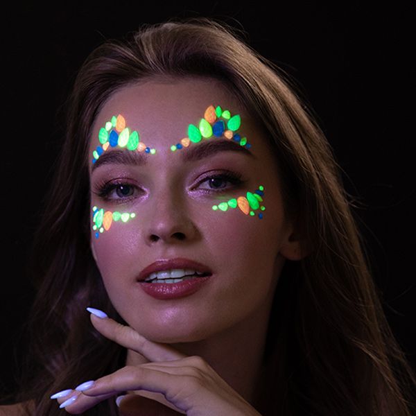 A person's face adorned with glowing, multi-colored self-adhesive face jewels on the forehead and cheekbones, emitting a vibrant green, orange, and blue light in a dark setting, showcasing their glow-in-the-dark effect.
