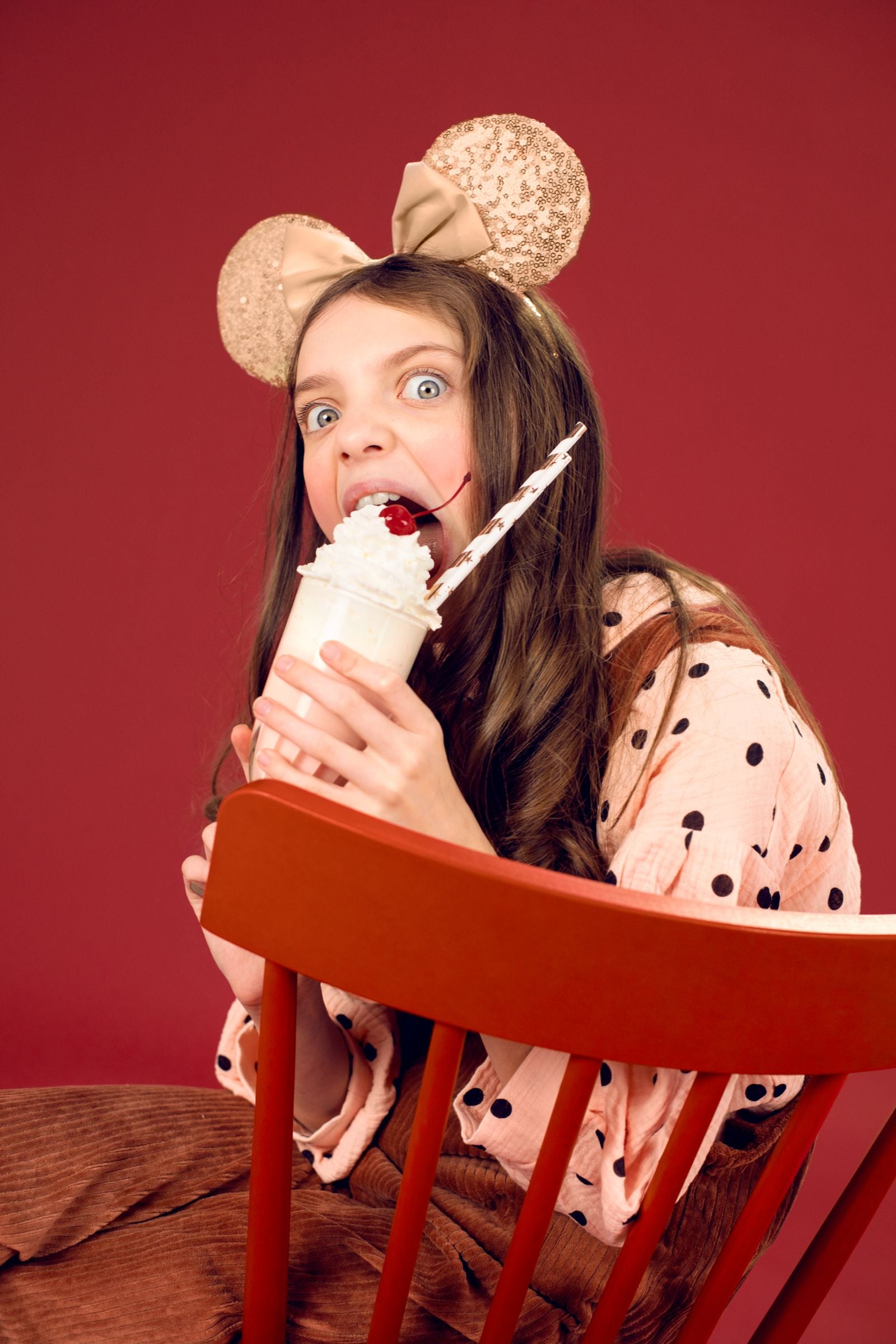 A person wearing a champagne gold sequined mouse ears headband, sitting backward on a red chair and playfully biting into a whipped cream milkshake with a cherry.