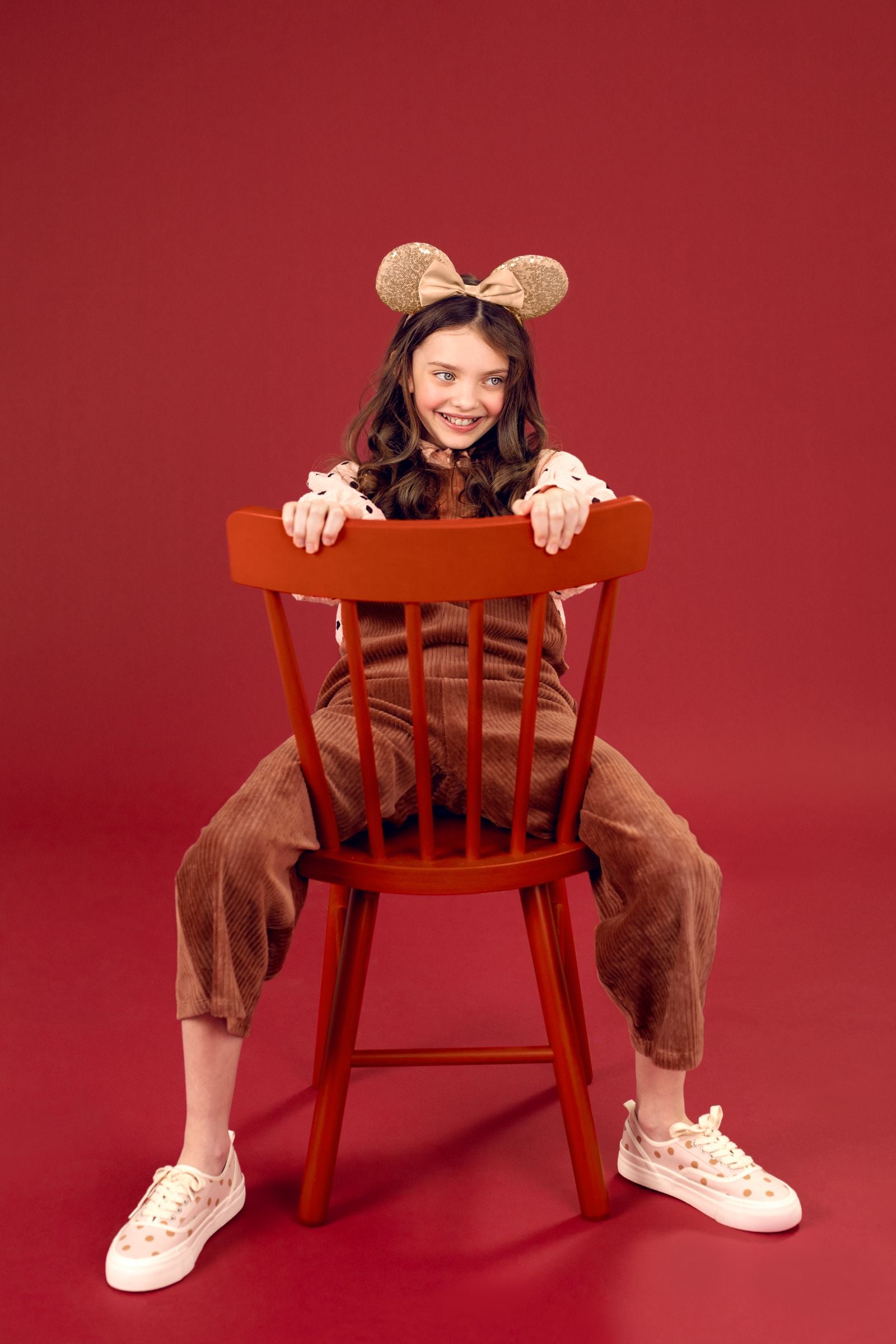 A person wearing a champagne gold sequined mouse ears headband, sitting backward on a red chair and playfully biting into a whipped cream milkshake with a cherry.