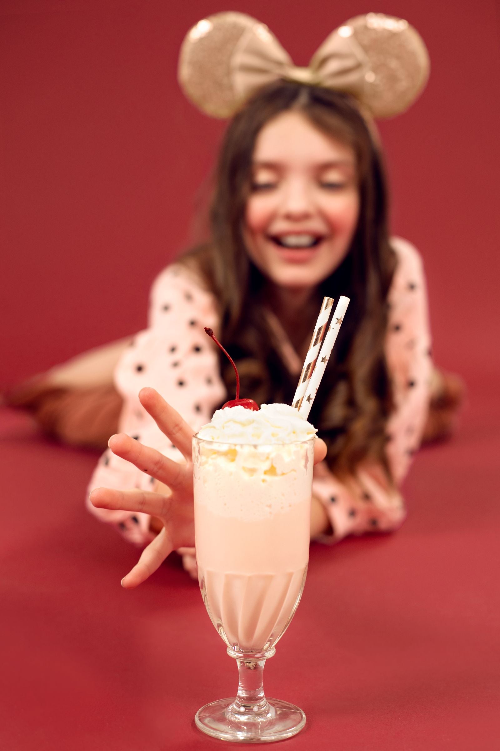 A close-up of a tall milkshake topped with whipped cream and a cherry, with a person wearing gold sequin mouse ears reaching for it in the blurred background.