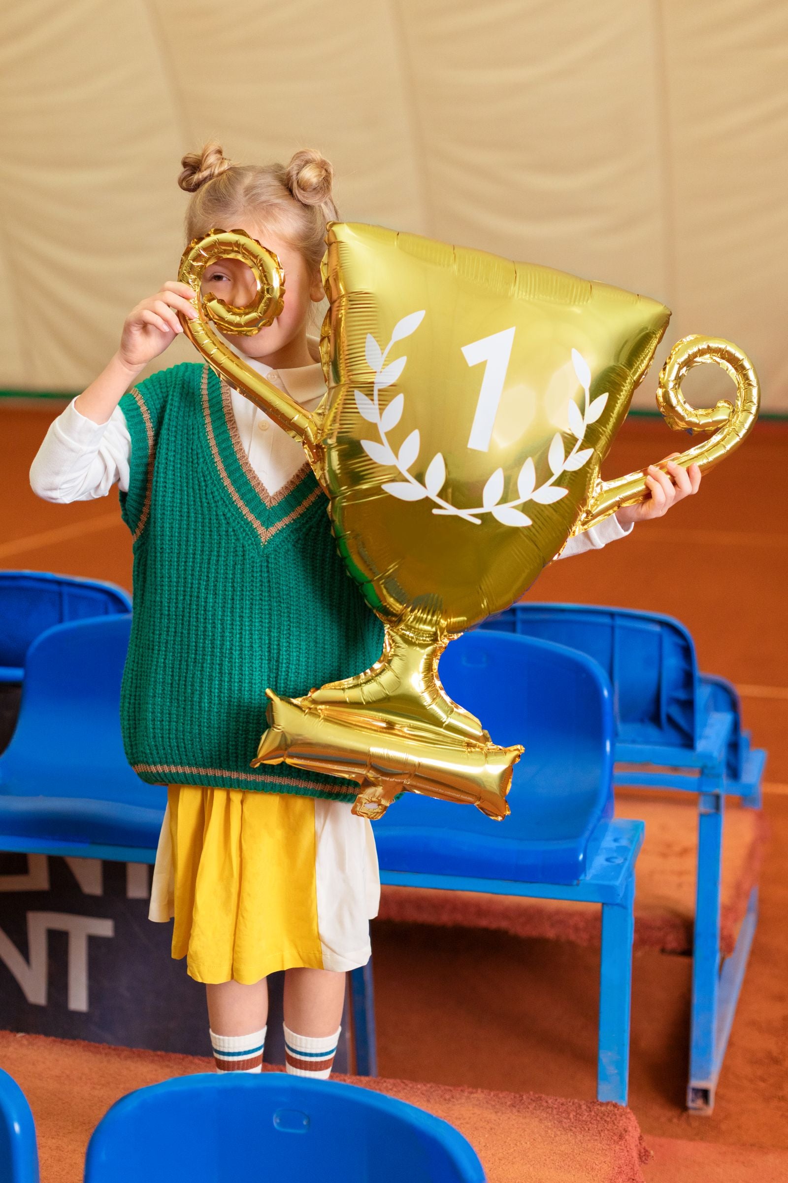Child holding a large gold trophy balloon with a number one on it, standing in a sports arena.