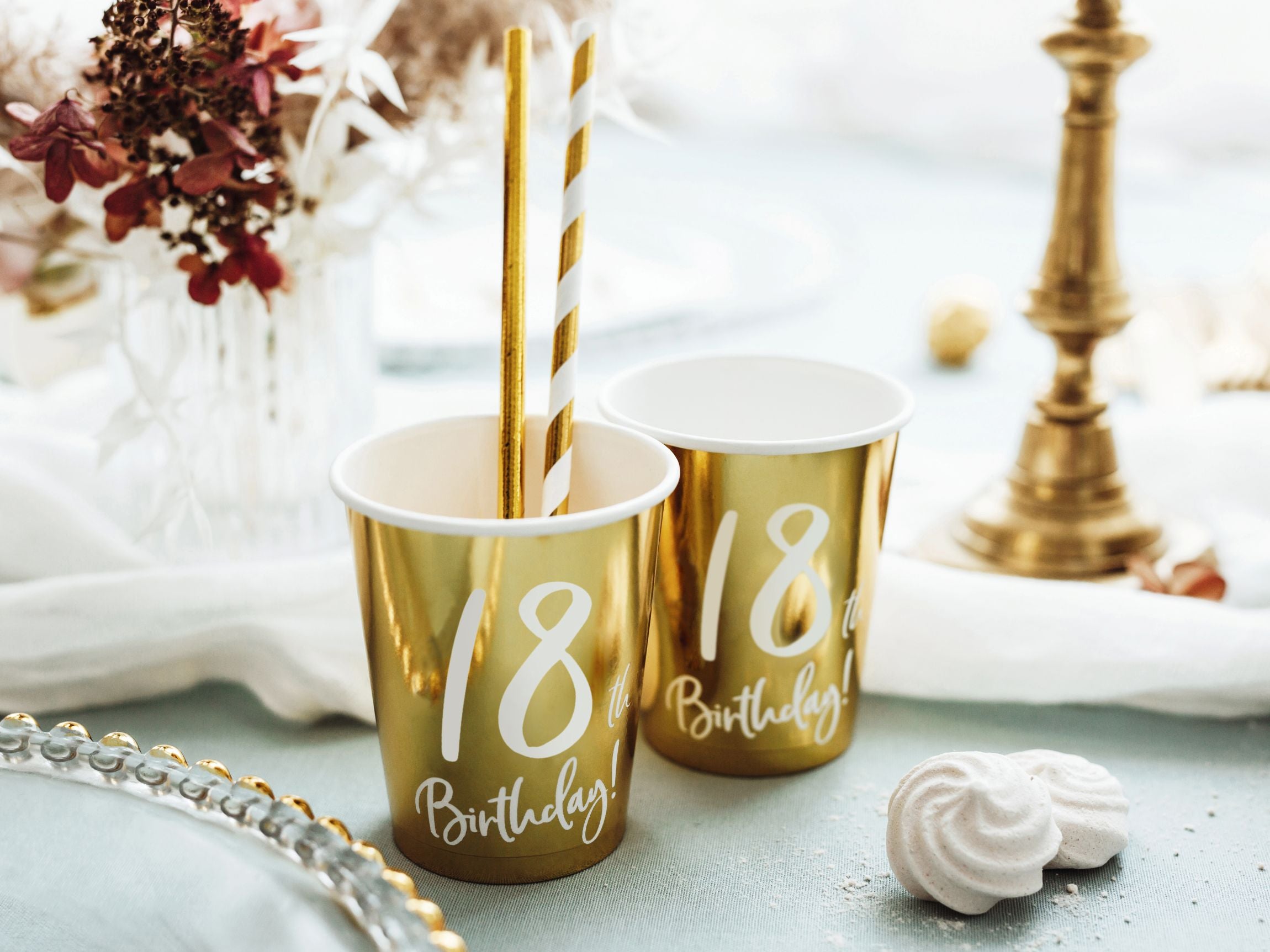 Two metallic gold paper cups with "18th Birthday!" written in white, each containing a gold and white striped straw. They are placed on a light blue table next to white meringues, with a blurred background of a flower arrangement and a gold candelabra.