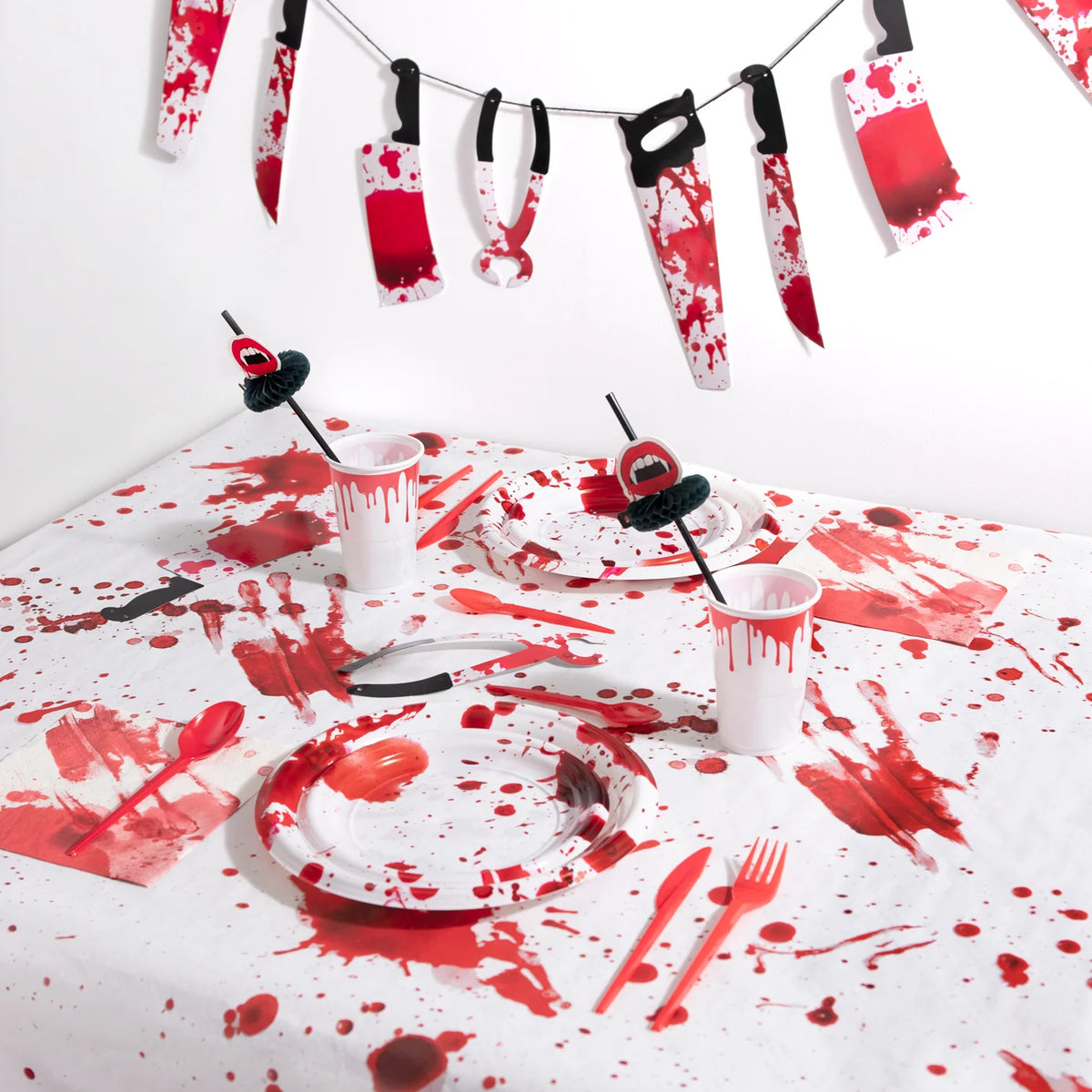A party table covered in a blood-splattered tablecloth, set with matching bloody plates and cups, sitting beneath a garland of paper weapons.