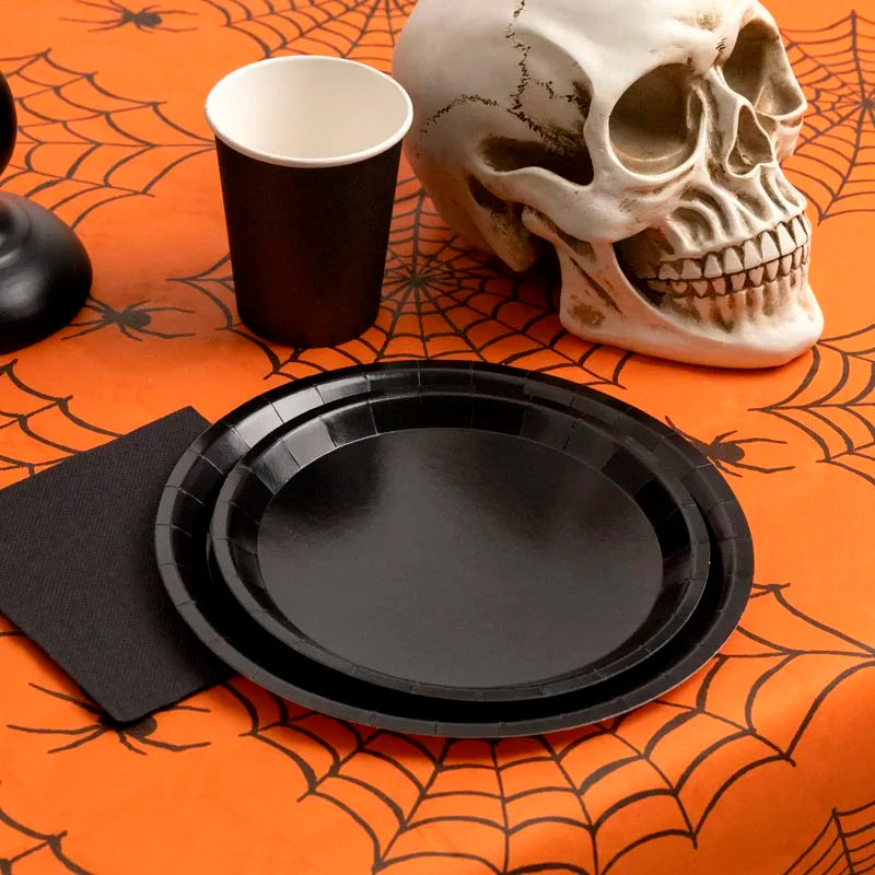 A decorative skull, a black paper cup, and black party plates sitting on a bright orange tablecloth featuring a black spiderweb print.